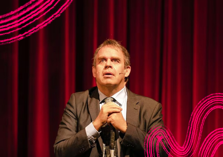 Performer Christopher Bailey. Christopher is dressed in a suit and looking upwards with his hand clasped in front of him. He stands in front of a red velvet curtain. Around him are bright pink swirls and wavy lines.