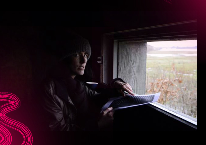 Artist Suzanne Walsh sits in front of the window of a bird hide. The room is black and the only light comes from the window, it barely illuminates her face. Through the window a marshy landscape is visible. Suzanne holds a list in her hand. The left of the image fades to black and against this are bright cerise pink painterly lines.