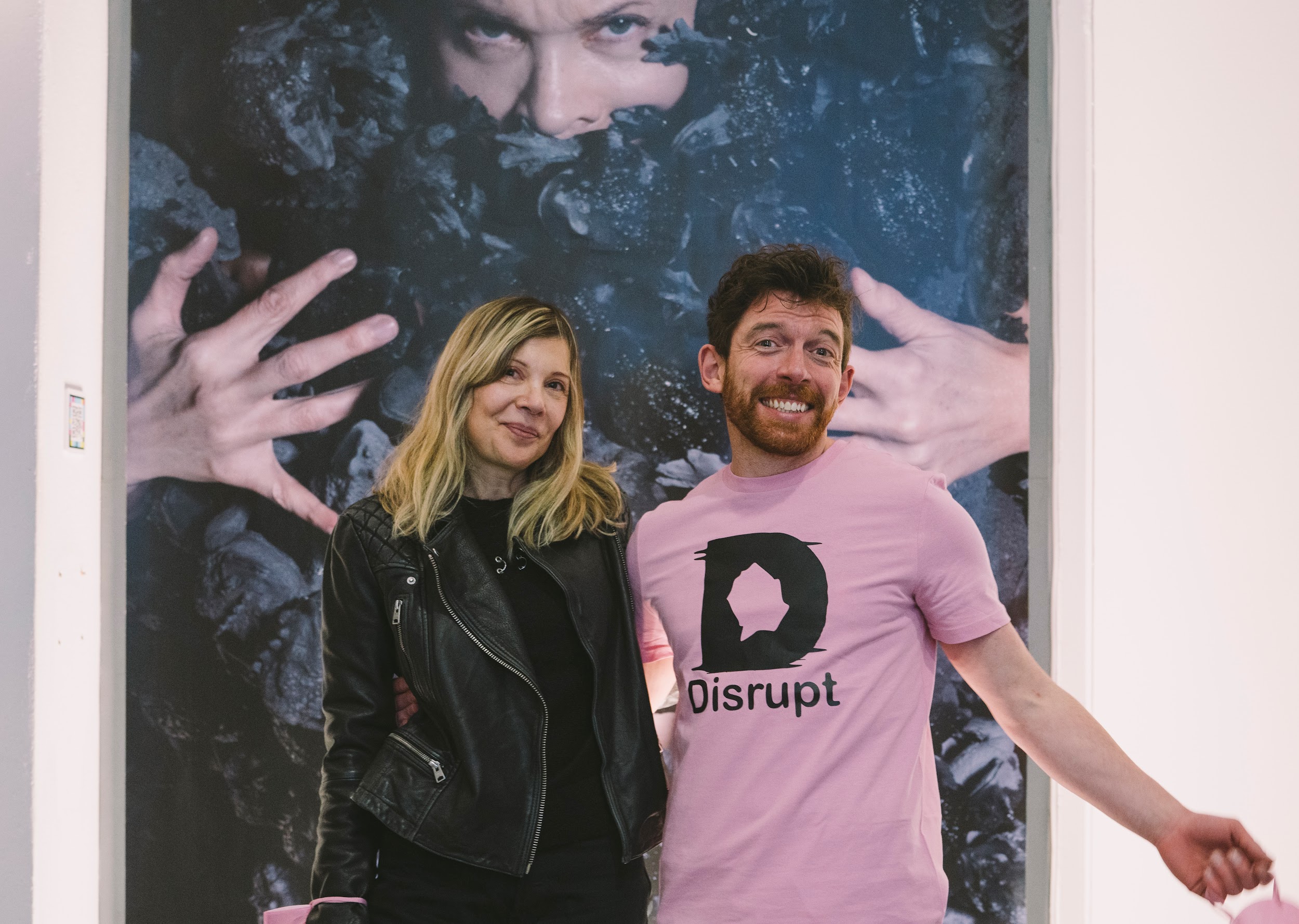 Visual artist Cecilia Bullo stands with Distinct curator Alan James Burns. Their arms are intertwined, both individuals are beaming. Behind them is an artwork of Cecilia’s. A large-scale photographic print. In the photograph is submerged in a bed of clay frogs. Photo by Leanne Sullivan.