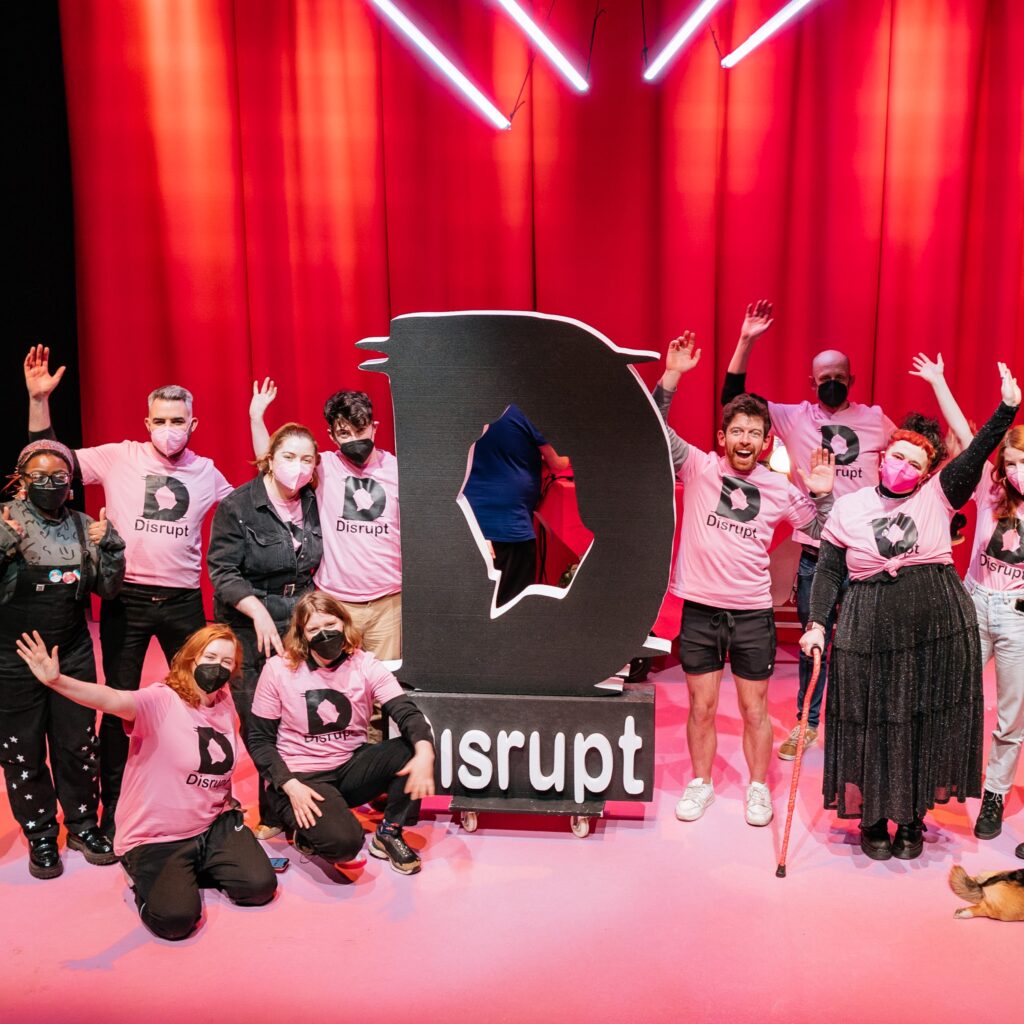 A group photo of 13 members of the Disrupt festival team on stage. They are wearing pink festival t-shirts printed with the Disrupt Logo. The stage is heavily clad in pink tones, a light pink carpet and hot pink curtains. They are also joined on stage by a corgi service dog and a giant foam cutout of the Disrupt Logo in black. Some strip lighting is hanging overhead. Photo by Simon Lazewski.