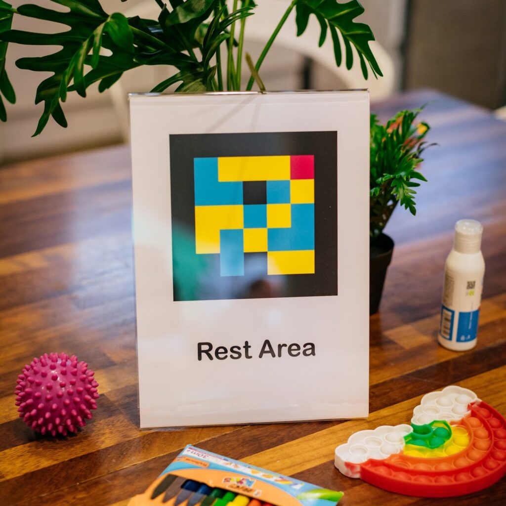 In the lobby of Project Art Center is a sign which is propped up on a table. The sign reads “Rest Area” it also has a Navicode image on it. Other objects on the table include. A plant, two bottles of hand sanitiser and two stim toys, one the shape of a rainbow, the other a pink ball.