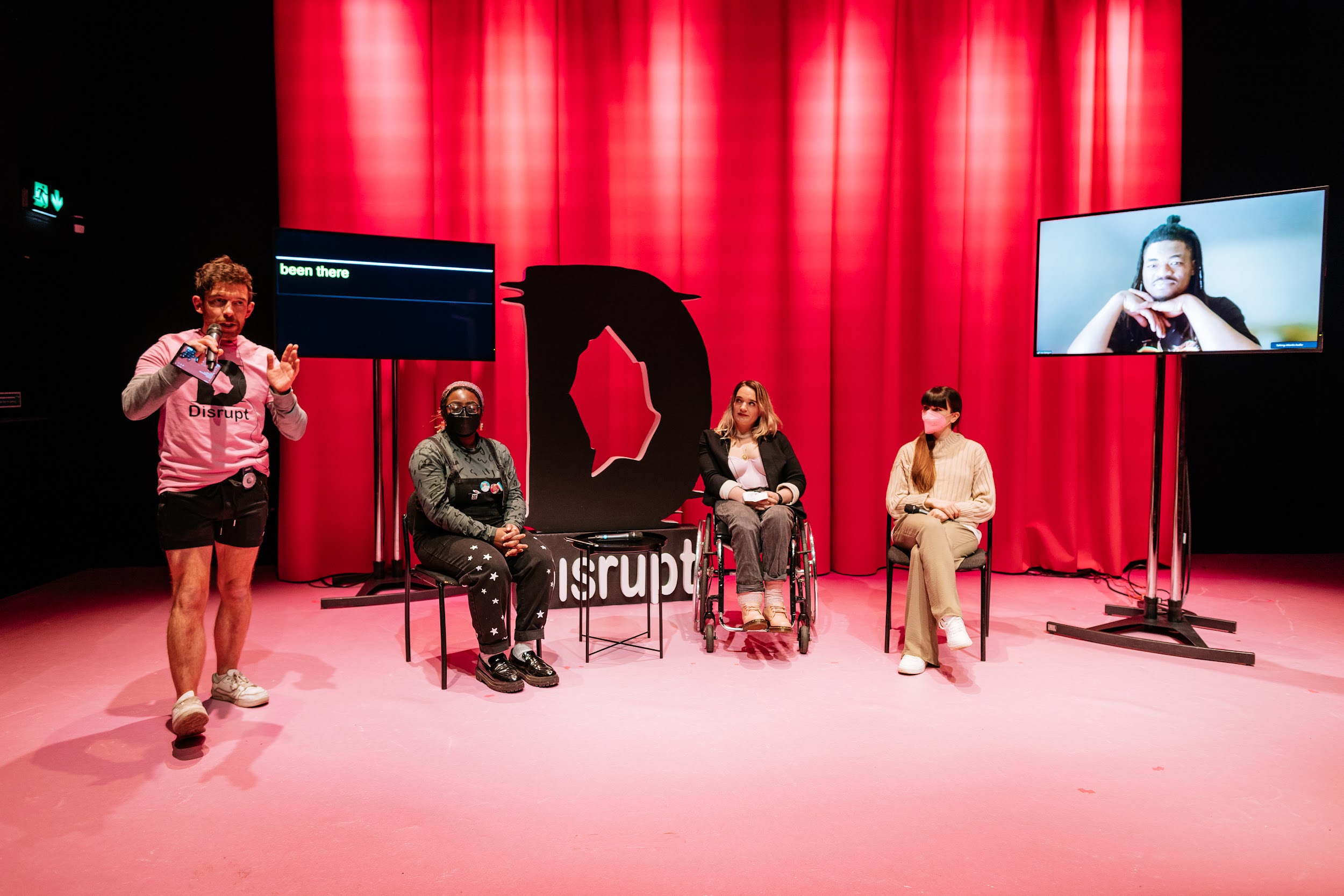 Disrupt festival organiser Alan James Burns stands on stage and addresses the crowd. Behind him are 4 Disrupt panellists. Three in person, One remotely, on a screen. Blezzing Dada who wears black and a face covering. Louise Bruton Who wears a satin corset and a black blazer, is seated in a wheelchair. Dancer Ali Clarke in an outfit of camel colours, with a pink face covering. On a large screen is Tobi Balogun. Tobi has locks and a topknot. Tobi’s elbows are propped up so that his head rests on his hands. Behind the group, and a screen shows captions of the conversation. Photo by Simon Lazewski.