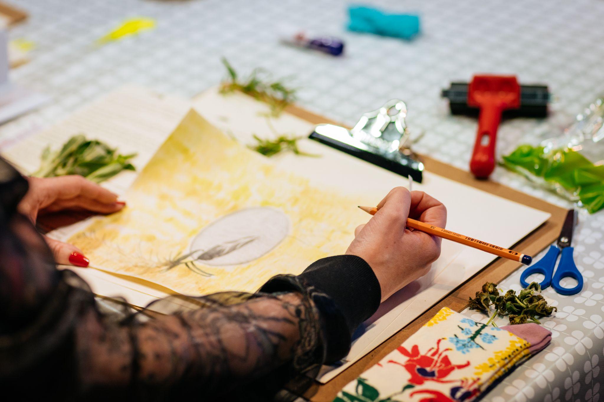 An over shoulder scene is captured. Here we have hands drawing a picture at a desk. They are wearing a long-sleeved chiffon-style black top and have brightly coloured red painted nails. On their table are various materials, including a clipboard with blank sheets, leaves, and scissors. Photo by Simon Lazewski.