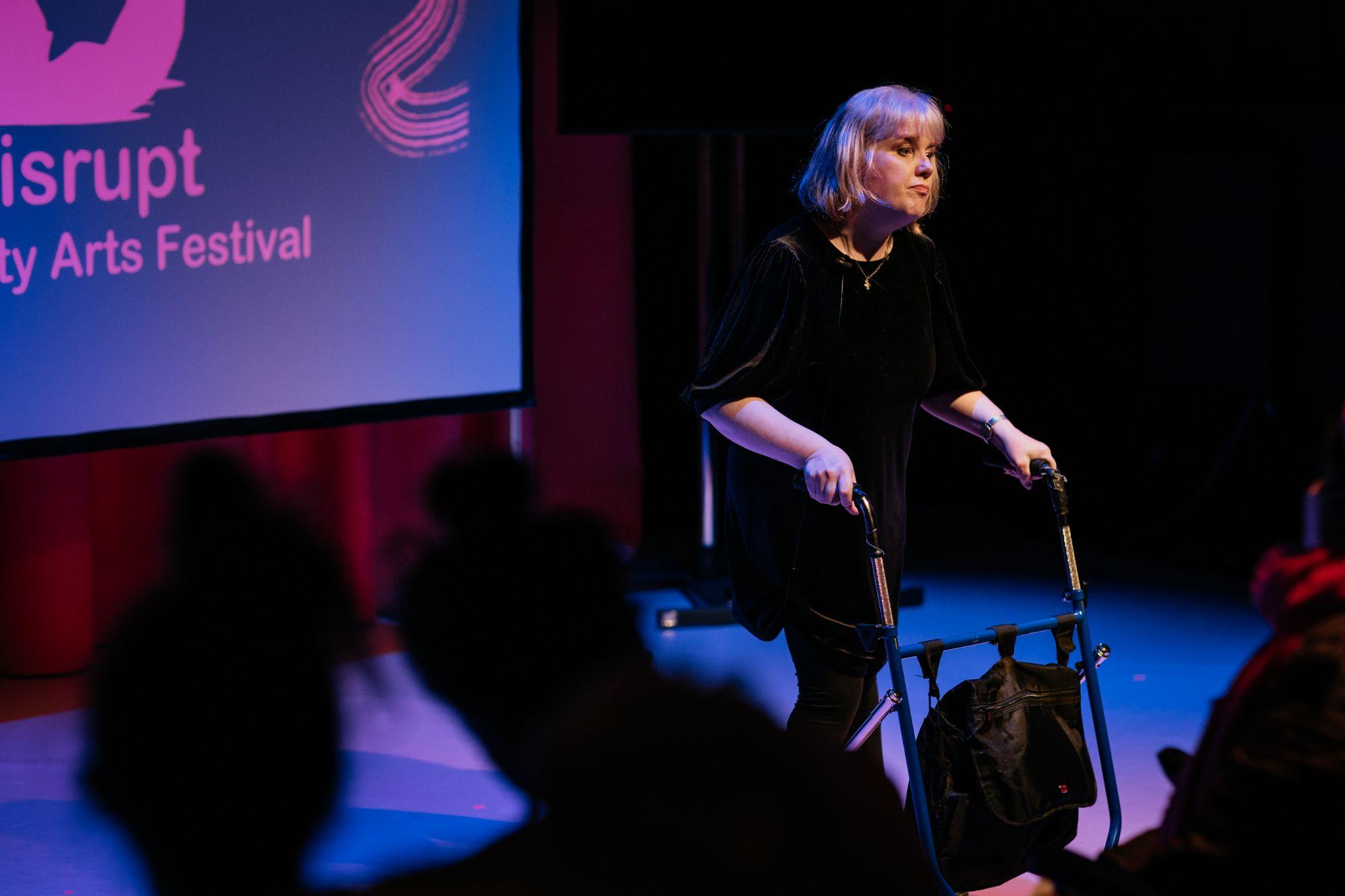 Artist Mairéad Folan stands on a stage lit in purple and pink hues. She is dressed all in black and leans on a black walking frame. They engage with an audience, who are mostly unseen. The stage is lit in purple and pink hues. Viewed from a diagonal perspective within the audience area, silhouetted spectators sit facing the stage. In the background there is a projection screen displaying the logo for the Disrupt Disability Arts Festival. Photo by Simon Lazewski.