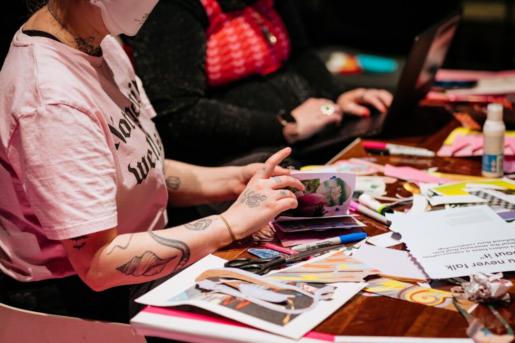 A figure wearing pink, is sitting at a desk which is covered in coloured paper, marker and magazine cuttings. They are making a zine. They have pale tattooed arms and wear a t-shirt with pink lettering. Photo by Simon Lazewski.