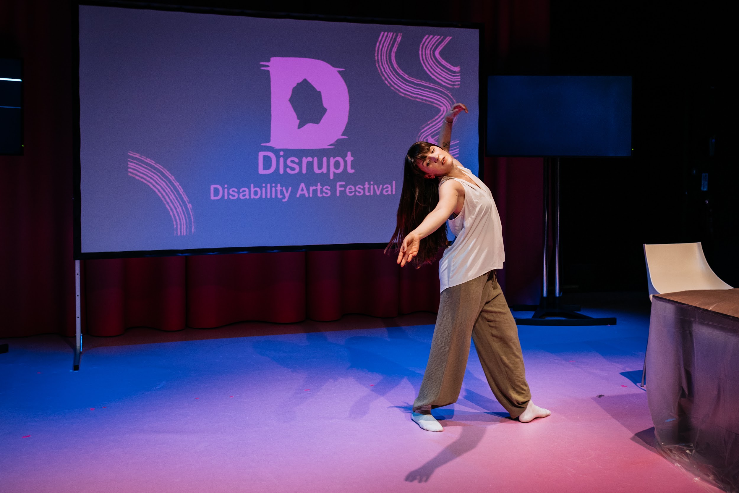 An image of dance artist Ali Clarke performing on stage. She stands in front of a purple background. Alison contorts her body, leaning deeply backwards in motion. She stands balancing, with one arm stretched outward as if trying to grasp something unseen. Photo by Simon Lazewski.