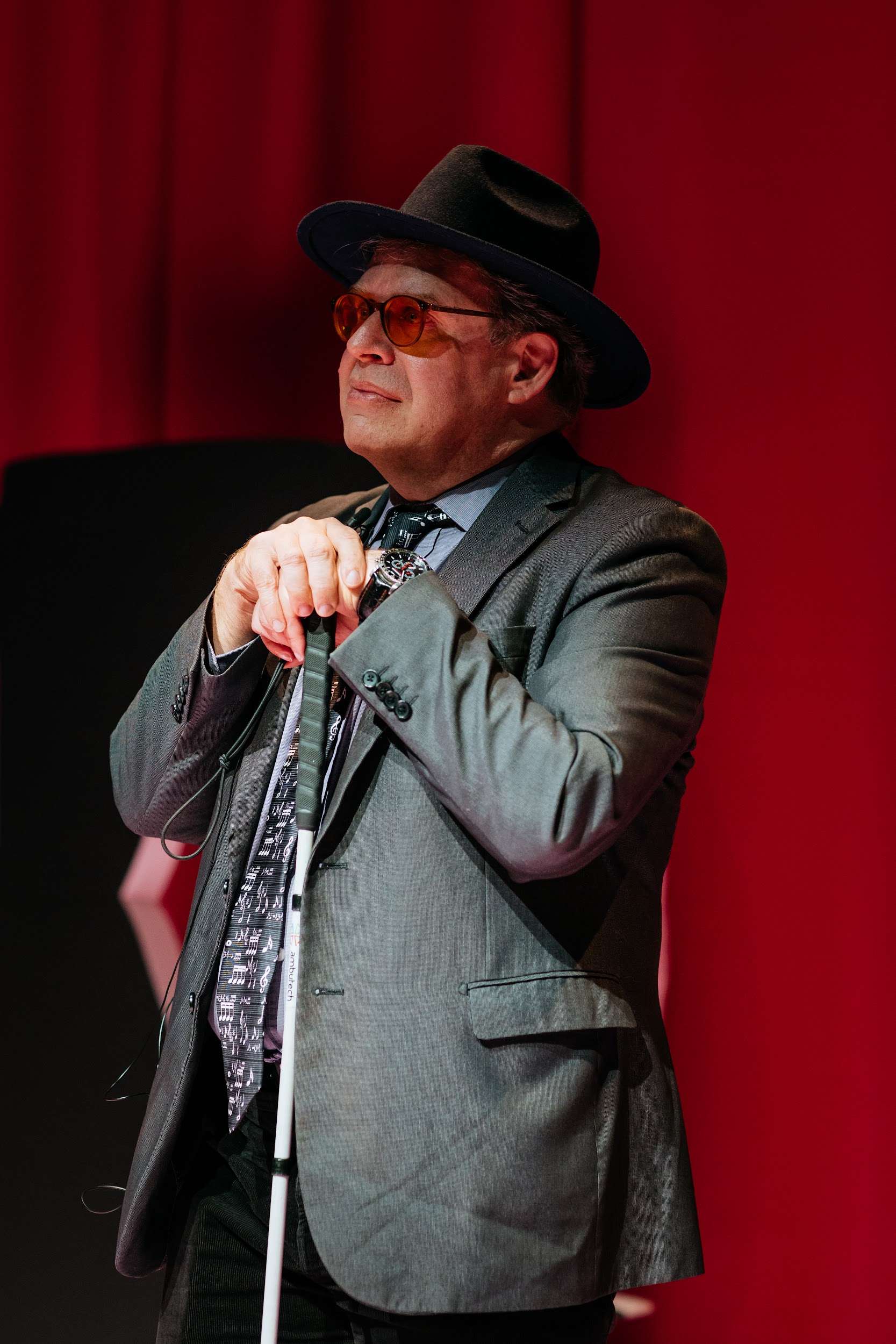 Artist Christopher Bailey stands in front of a pink curtain. He wears a suit jacket, shirt, tie and wide brimmed hat. He leans on a cane, gazing outward in a confident and content manner. Photo by Simon Lazewski.