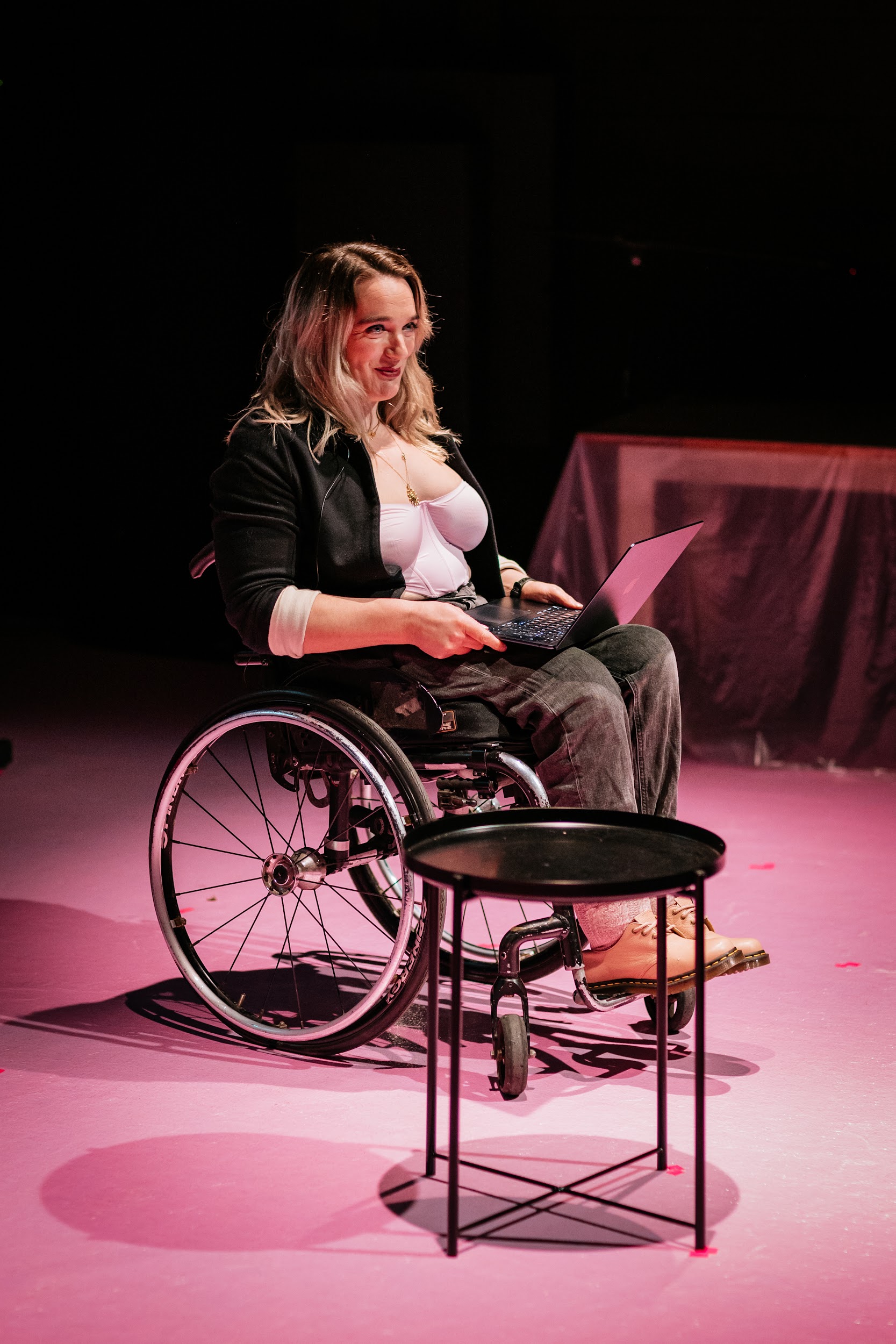 Writer Louise Bruton on stage. Lit in Pink Hues. Louise has tumbling blond curls. She wears black jeans, a black blazer and a pink satin corset. She is seated in a wheelchair with a laptop on her knee. On her face is a deep, warm expression. Photo by Simon Lazewski.