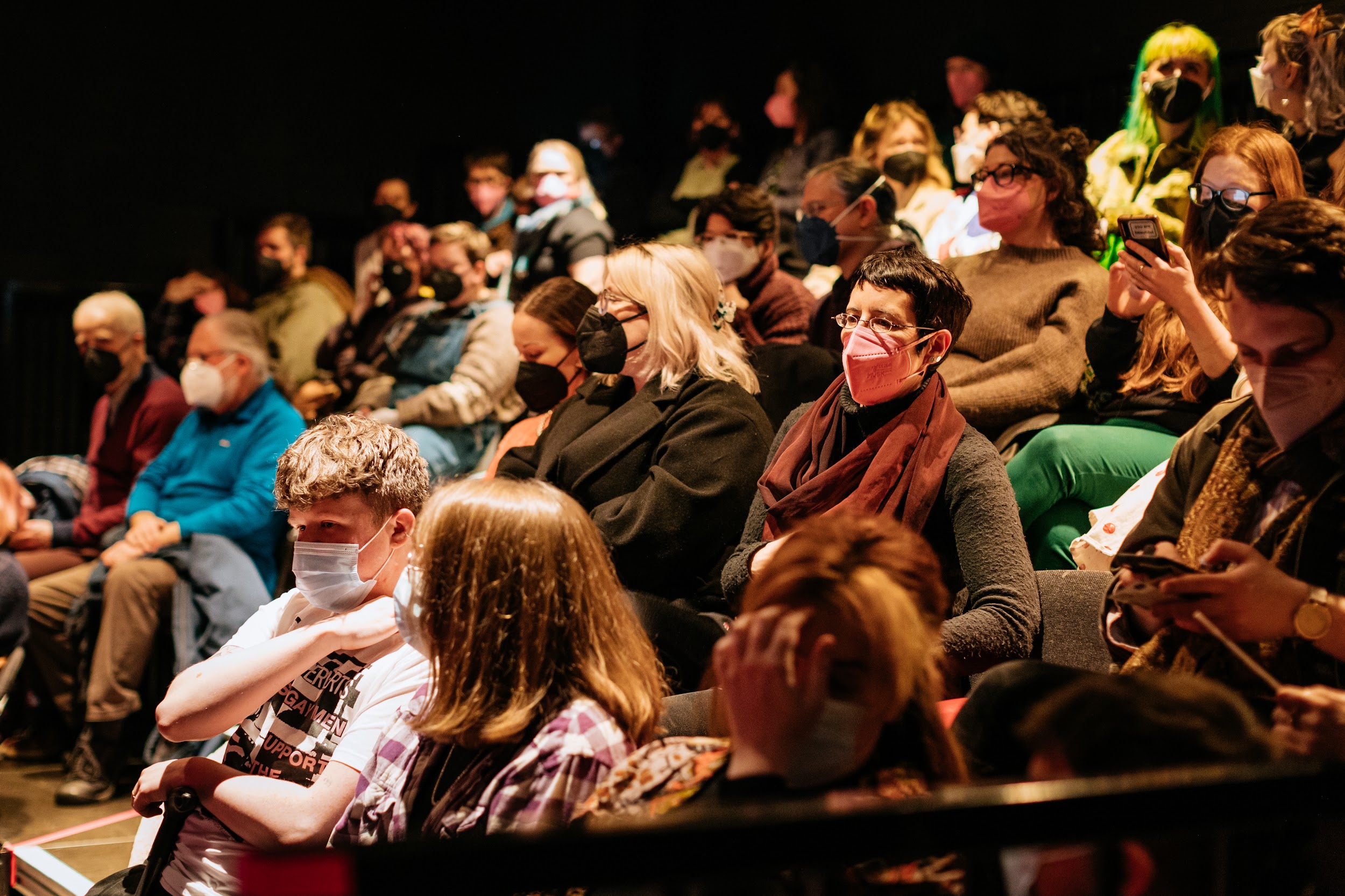 A crowd sit in tiered seating. They tentatively await a performance. The house lights are up. Everybody wears face masks. Photo by Simon Lazewski.