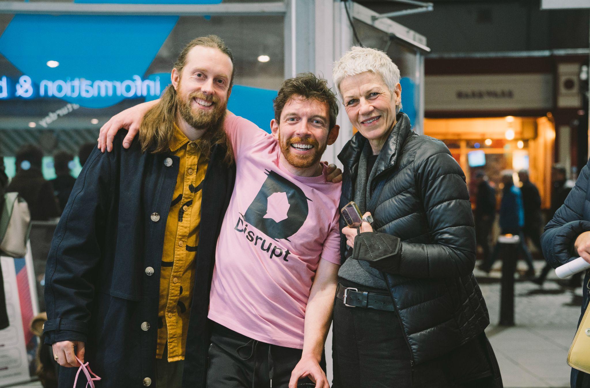 The image shows artist and curator AlanJames Burns standing with two people on either side, all facing the camera and smiling with arms around each other. Alan is dressed in a pink t-shirt featuring the Disrupt Disability Arts Festival logo. In the background and out of focus, an open door reveals a busy city street at night. Photo by Leanne Sullivan.