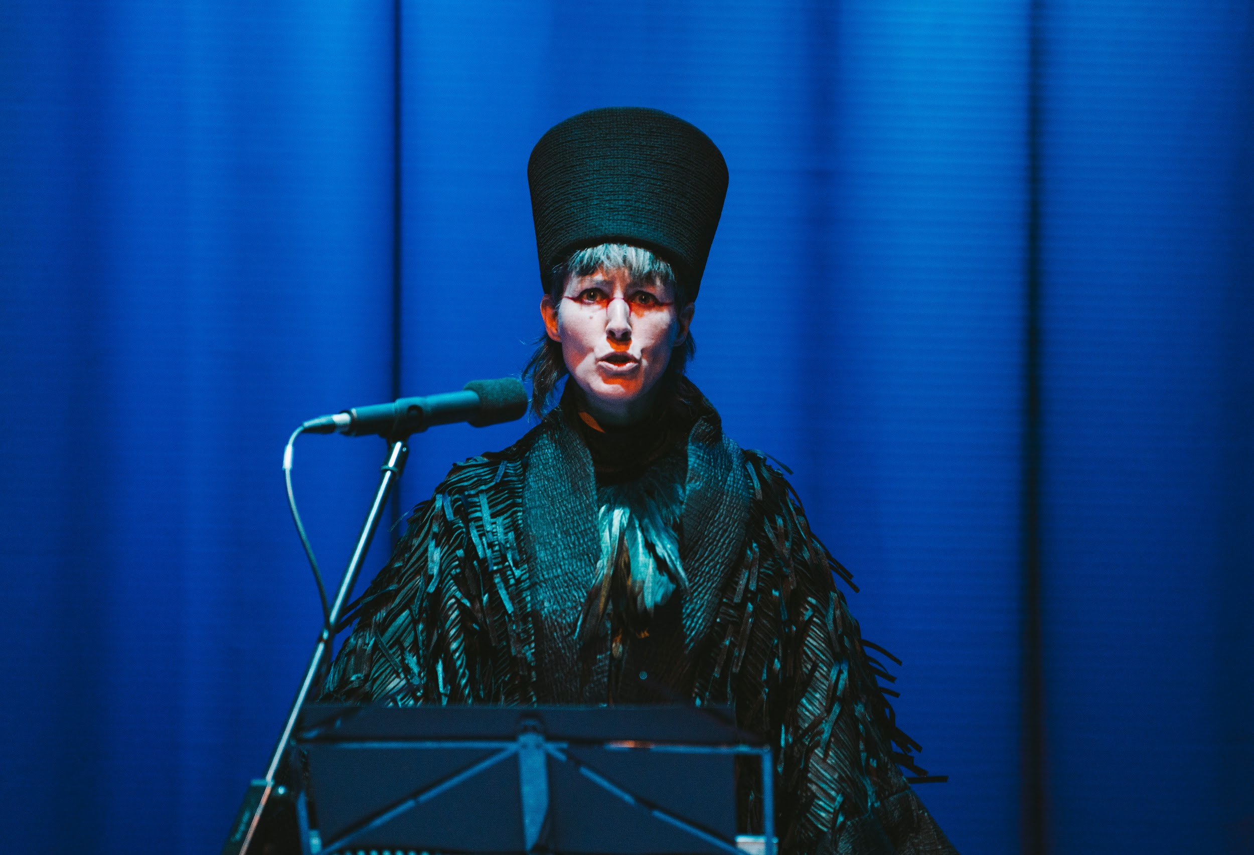 Performance artist Suzanne Walshe on stage in action. They are standing in blue lighting. They wear ceremonial dress, a ruffled black cloak, birdlike. A tall head dress . Suzanne speaks dramatically into a microphone. Photo by Leanne Sullivan.