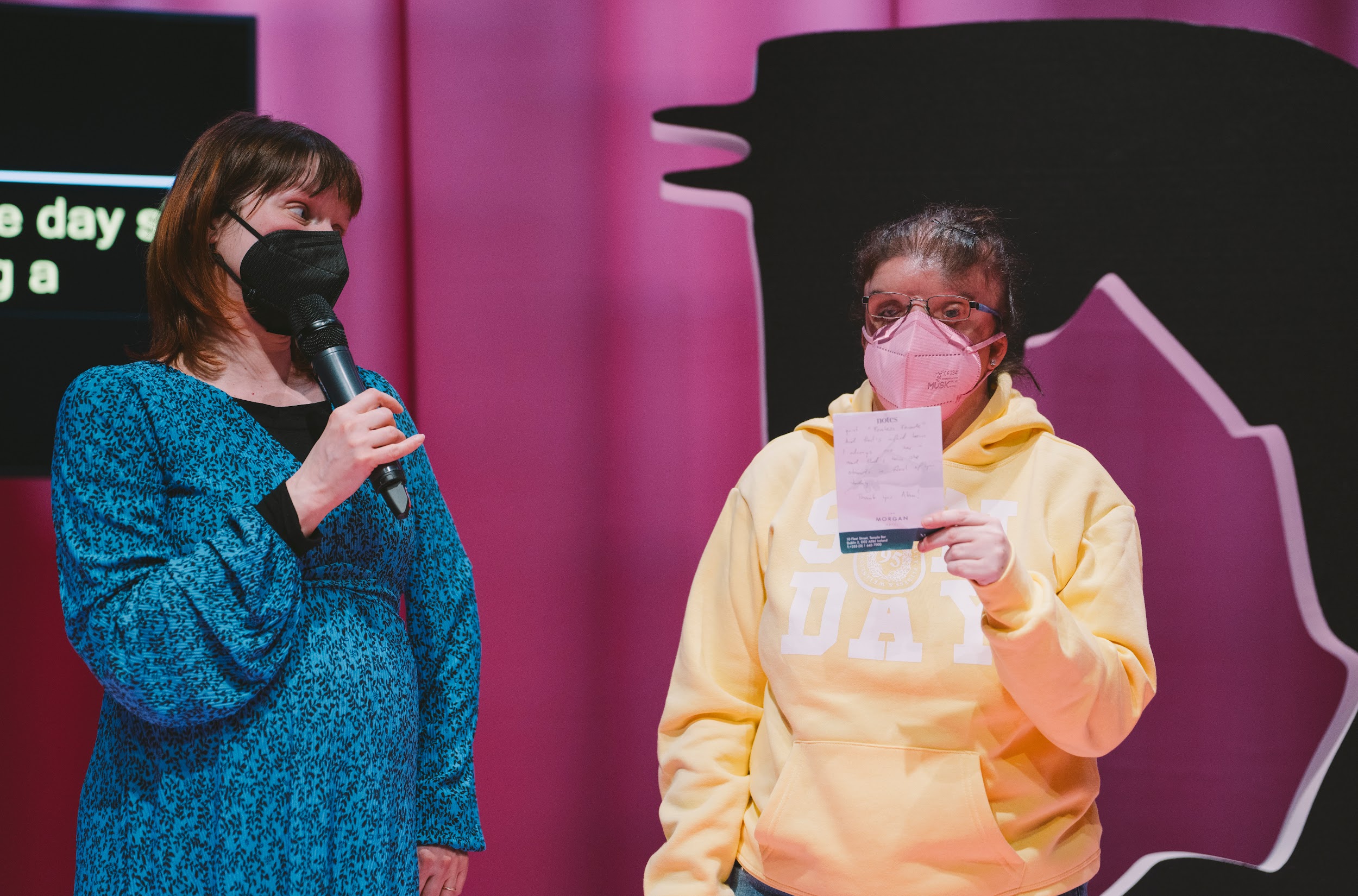 Two feminine figures stand on a stage. One, in blue wearing a face mask looks to the other, while speaking into a microphone. The other person, artist Yvonne Condon looks outward. They are wearing a yellow hoodie. Behind Yvonne is a large styrofoam D. The artist Yvonne holds up a small white piece of paper. Photo by Leanne Sullivan.