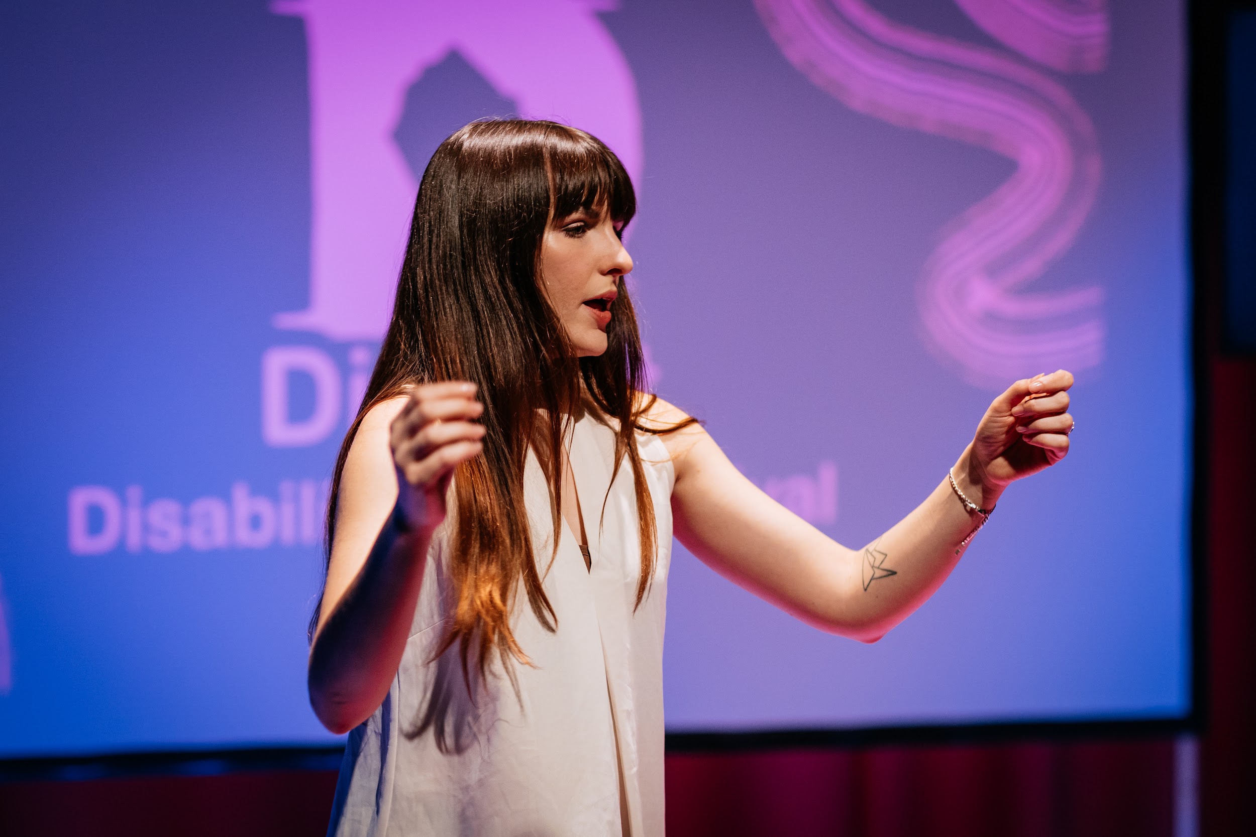 An image of artist Ali Clarke performing. Ali is turned slightly away from the camera, and we get her side profile. She is a white woman with long brown hair with a fringe. She wears a white sleeveless blouse and has her two arms outstretched in motion. She is well lit and behind her we see a projection of the Disrupt logo on a screen with pink and red hues. Photo by Simon Lazewski.