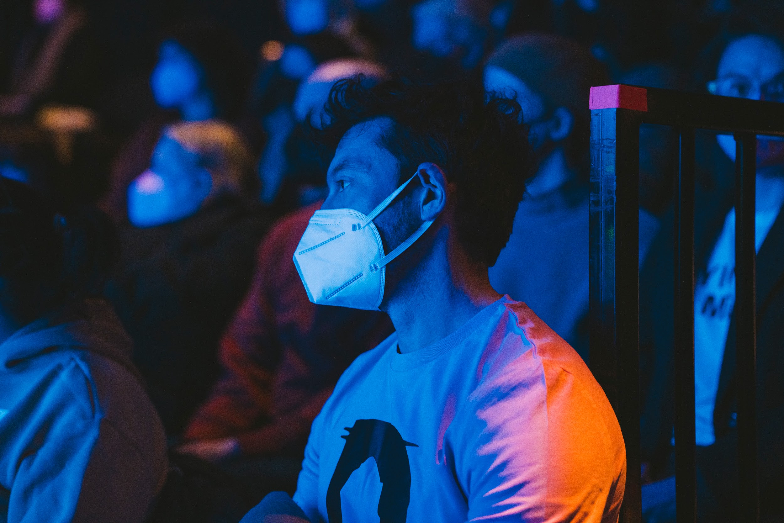 Disrupt festival organiser, sits amongst a crowd. They wear a pink t-shirt and face-mask. They gaze toward a stage, with an expression of delight. They are lit in hues of blue. Photo by Leanne Sullivan.