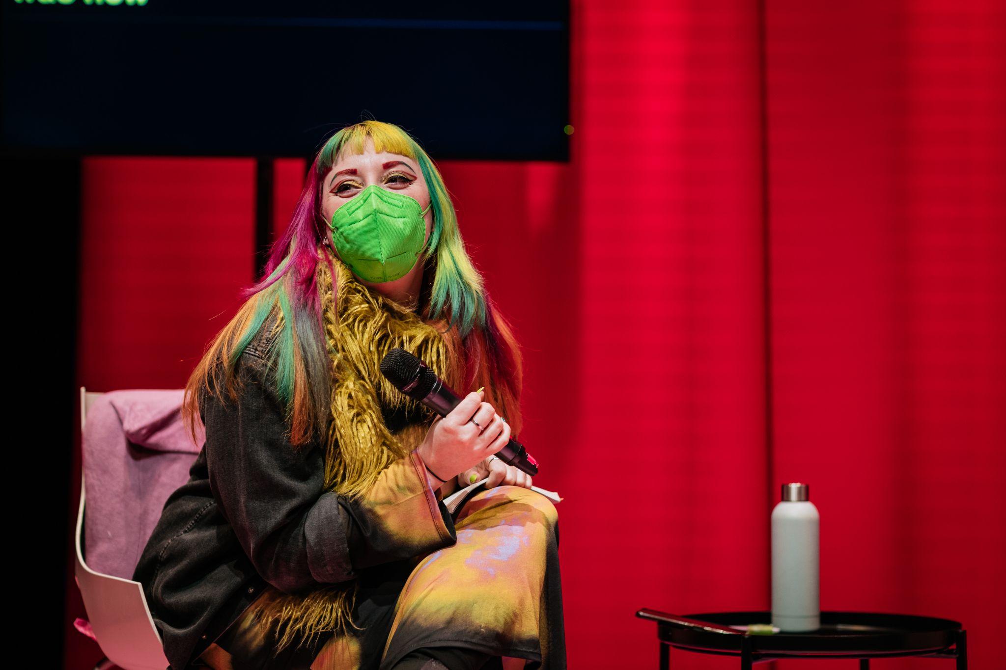 Tara Carrol sits on a chair at a slight angle to the camera, with their head turned to their right. They have long multi-coloured dyed hair and wear a bright green mask, a black jacket with yellow fur, and are holding a microphone in their right hand. A small black table to the right of the frame holds a water bottle. Behind them is a vibrant pink background, with a black monitor on a stand mostly out of frame. Photo by Simon Lazewski.