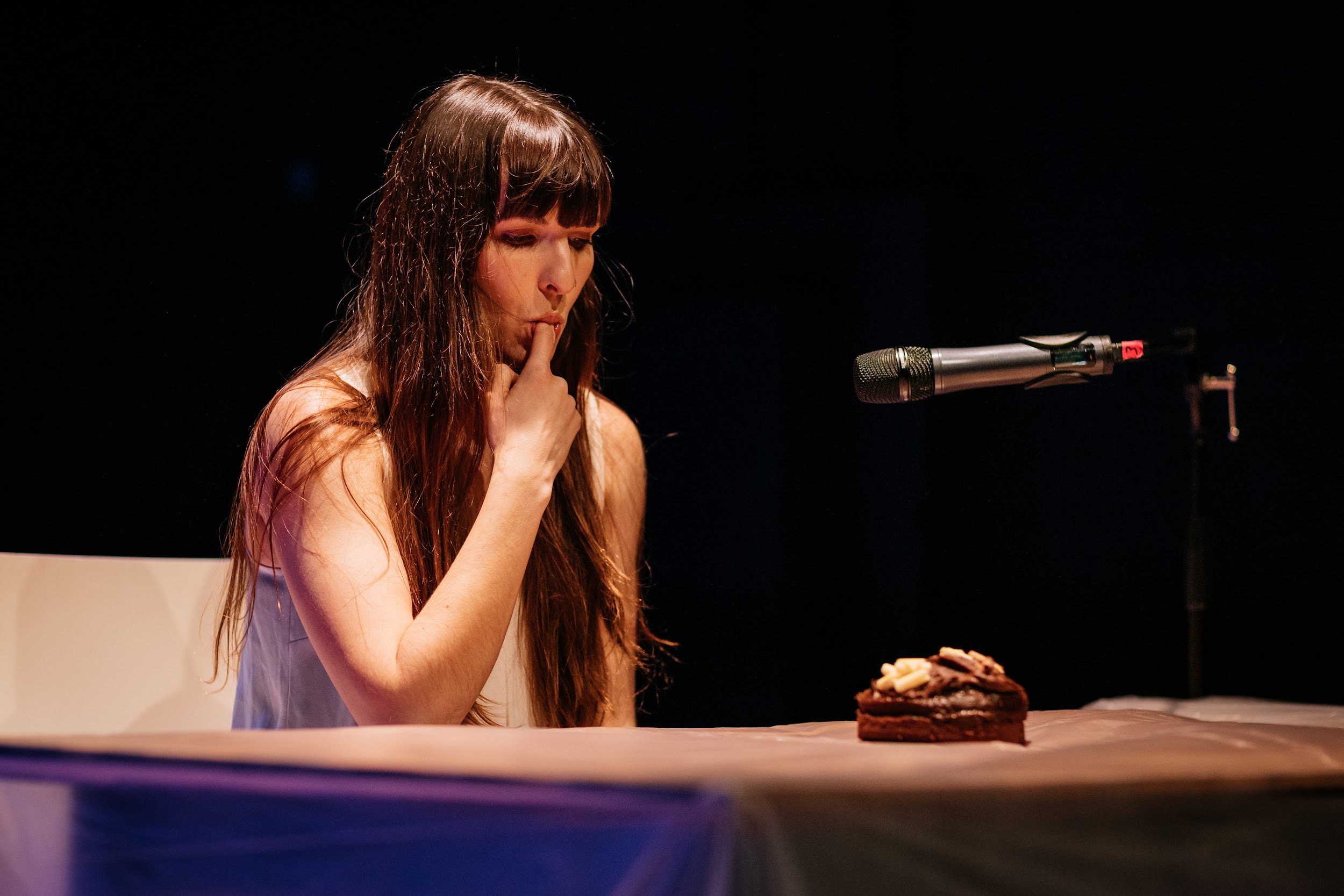 Dancer Aisling Reina is pictured sitting at a desk in stage lighting. A microphone nearby. Upon the table is a small chocolate cake. Alison, who wears her long, straight dark hair down, licks her index finger. It is a quiet moment. Photo by Simon Lazewski.