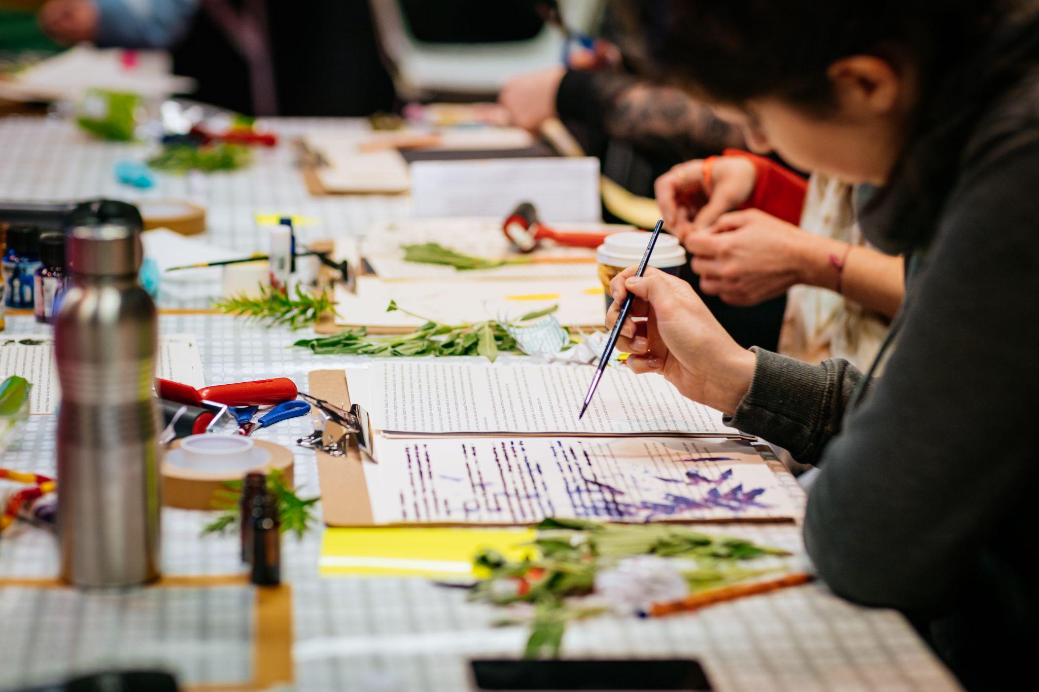 An image taken from the side shows people sitting at a long desk engaged in art and craft activities. The person in the foreground appears to be painting onto a sheet covered in texts, while those behind her are out of focus. Various materials, including more texts, leaves, and paints, are on the table. Photo by Simon Lazewski.