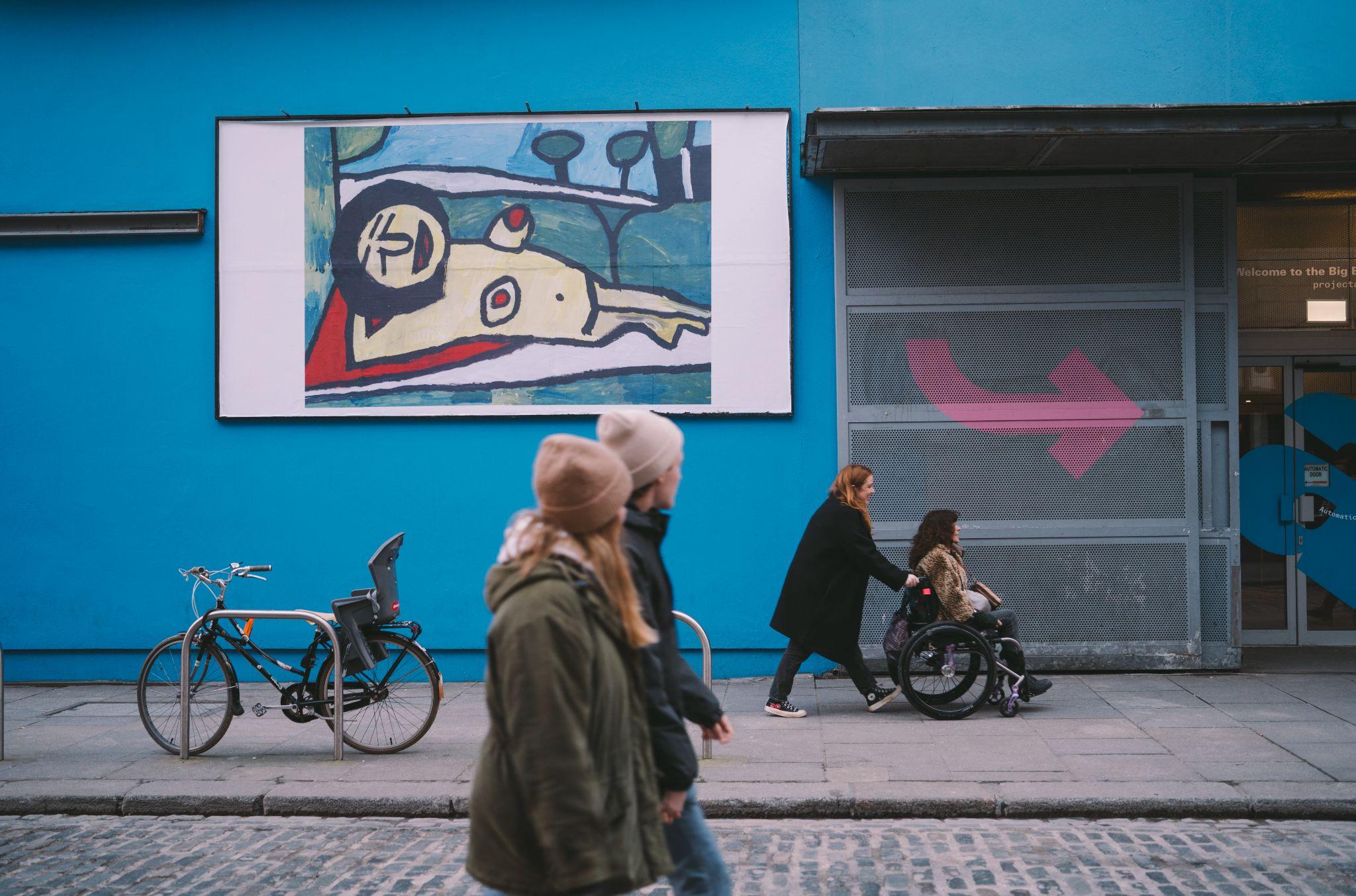 A billboard outside Project Arts Centre in Dublin, Ireland, displaying 'Yellow Nude' by Yvonne Condon against a bright blue wall. Taken from across the street, the image also features people walking in the same direction on both sides, including one person in a wheelchair. A bike is attached to a metal railing below the billboard on the left, while the entrance door to the centre is visible on the right. Photo by Leanne Sullivan.