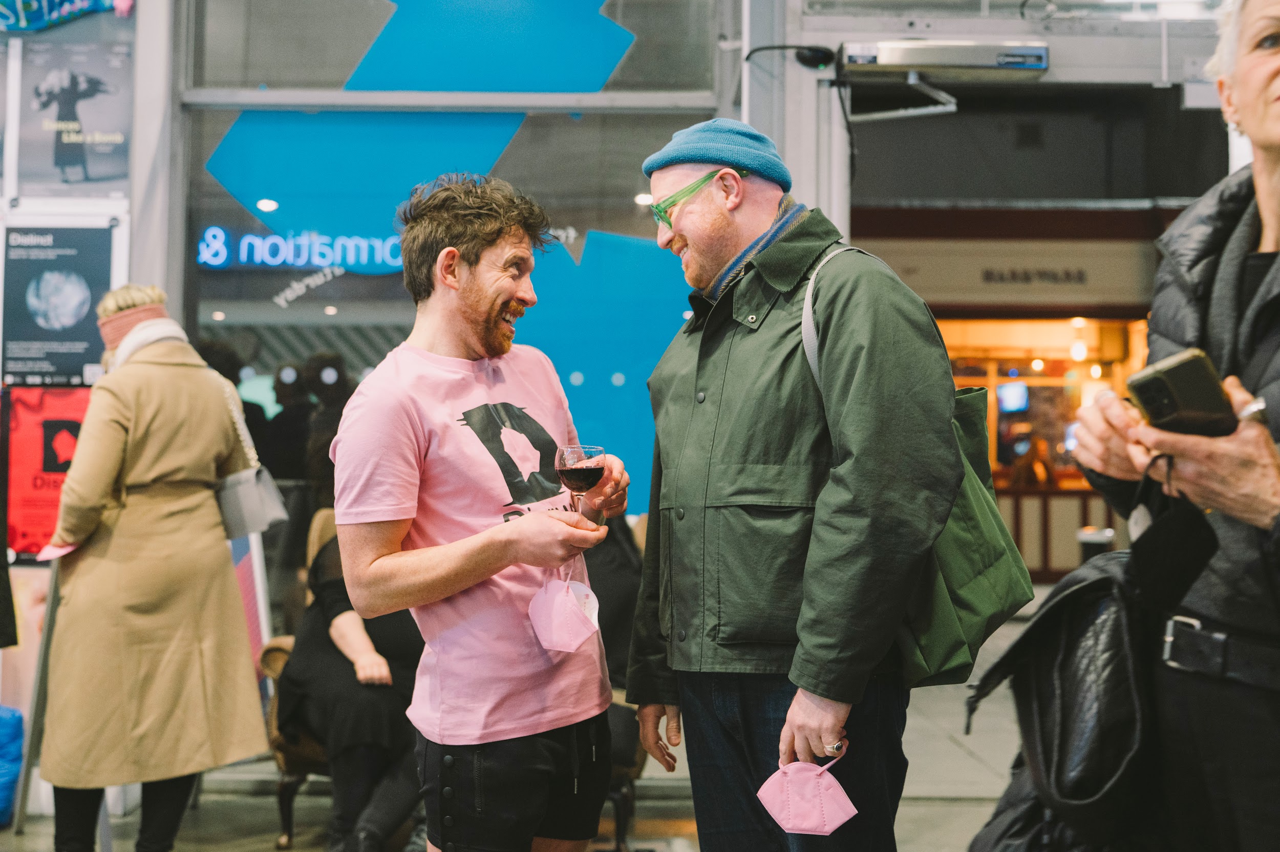 In a busy lobby artist and curator Alan James Burns socialises with the artistic director of Project Art Centre. Alan wears a pink Disrupt t-shirt. Cian wears a green jacket and glasses and a cyan hat. Both individuals are laughing. Photo by Leanne Sullivan. March 8th