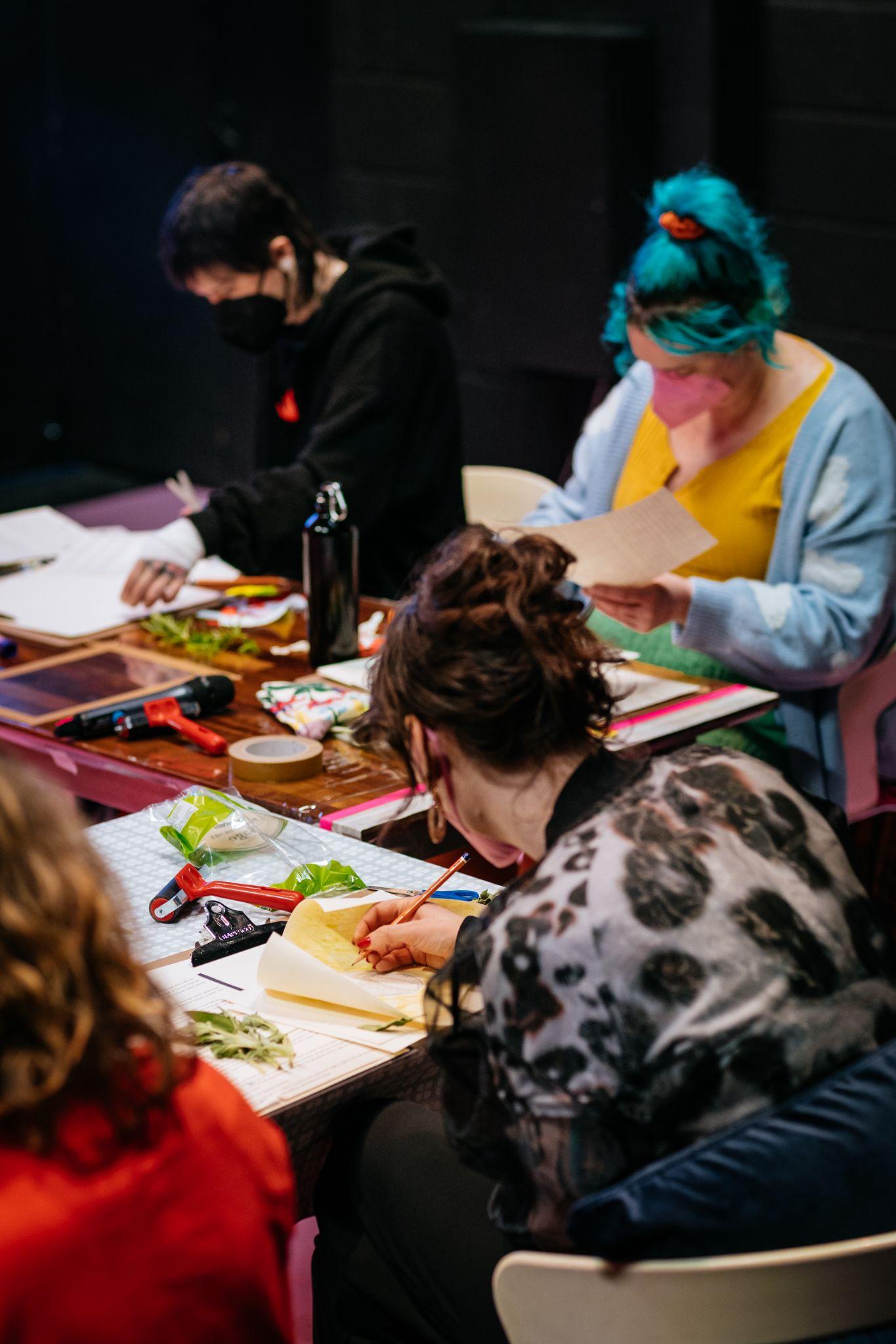 An image taken diagonally shows four people sitting at two desks positioned at an angle to each other in a workshop setting. They are all dressed in various colours and have different hair shades, one of which is dyed bright blue. They appear to be cutting paper with scissors and drawing. Various materials, including more texts, small boxes, and paints, are on the table in front of them. Photo by Simon Lazewski.