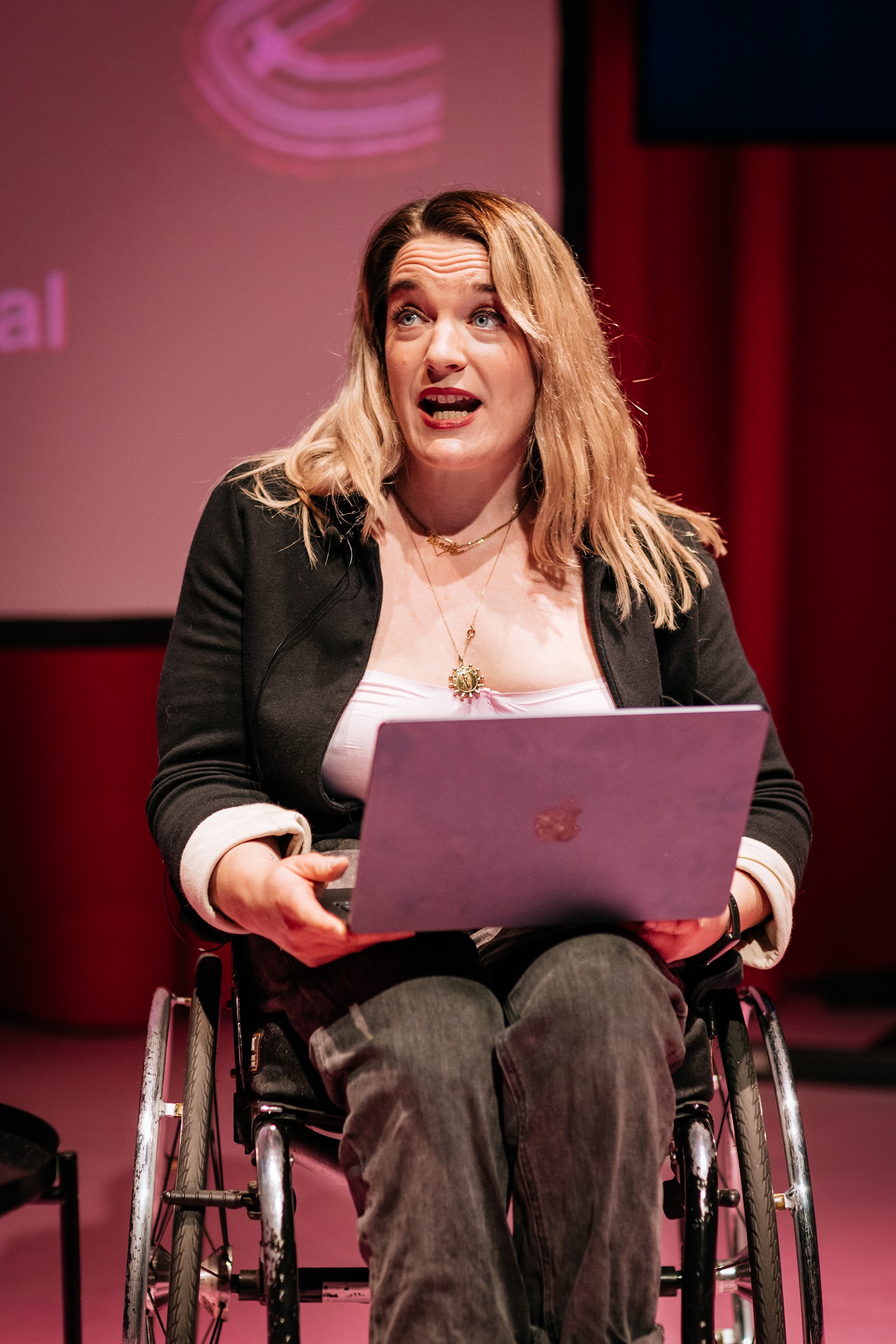 A portrait image of artist Louise Bruton performing. Louise is sitting in a wheelchair in the centre of the photo on stage, she has a laptop on her lap and is looking upwards towards the audience in conversation. She is a white woman with long blonde hair and wears a pink top and a black blazer. The angle of the photo is straight on and close up. The floor of the stage is a bright baby pink and is lit with pink hues. In the background we see a curtained dark pink backdrop and partially a projection screen. Photo by Simon Lazewski.