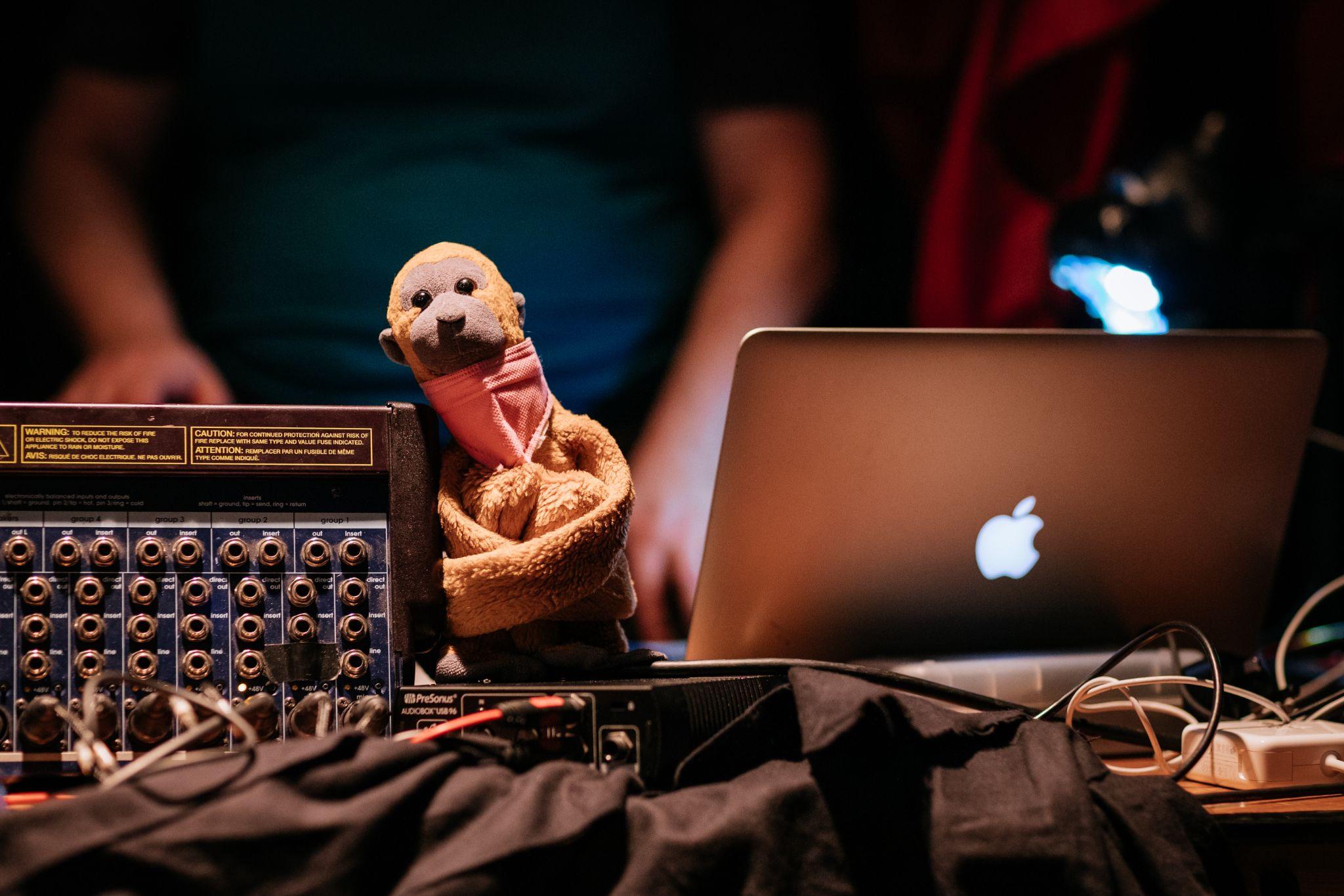 A small monkey-like teddy bear is positioned against technical equipment, facing the camera. In the background to the right, a MacBook emits light toward an out-of-focus figure. Wires and laptop chargers are visible on a wooden table, which is partially covered in black material in the foreground. Photo by Simon Lazewski.