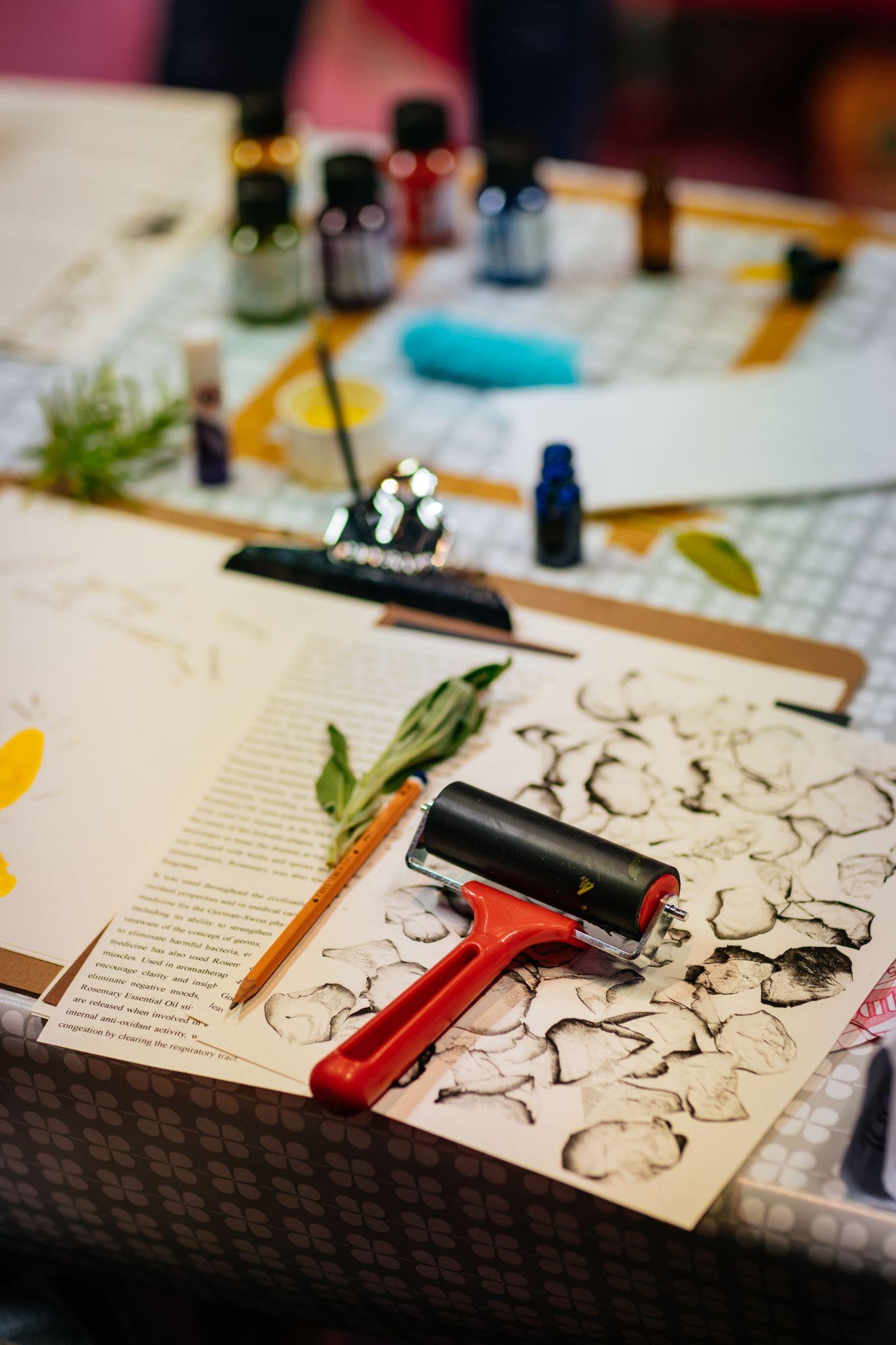 A desk with various art materials. This includes a brayer roller for printmaking, loose leaves, a white sheet with printed black leaves upon it, paper with text on, a clipboard, and various coloured paints. Photo by Simon Lazewski.