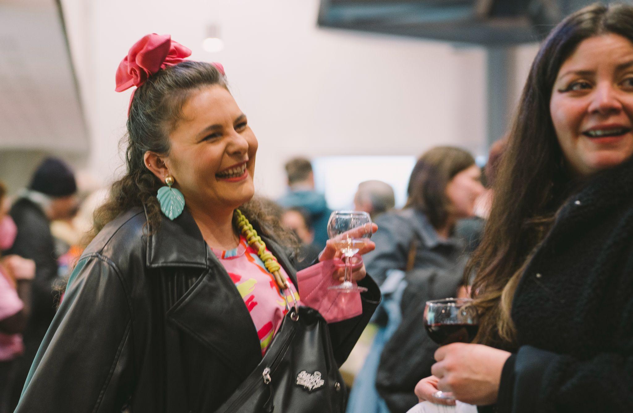 Two people with long dark hair stand in the foreground of the image, looking diagonally out of the frame and smiling. The person on the left wears a bright pink hair bow and a pink top. They both wear dark jackets and are each holding a glass of wine. Behind them, a large group of people is out of focus. Photo by Leanne Sullivan.