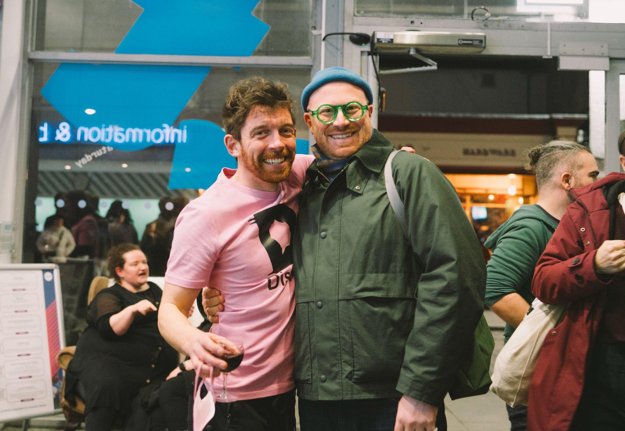 The image shows artist and curator AlanJames Burns and Project Arts Centre director Cian O’Brien, facing the camera and smiling with arms around each other. Alan is wearing a pink t-shirt featuring the Disrupt Disability Arts Festival logo, while Cian wears a dark green jacket and a blue beanie hat. Both individuals have redish hair and pale skin. The background is out of focus and reveals a lively scene with various people socialising. Photo by Leanne Sullivan.