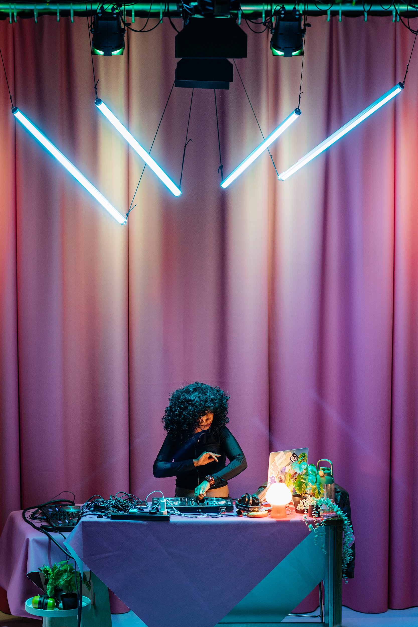 DJ Renn Miano depicted in portrait orientation, mid set. Renn wears black lycra and has voluminous black curls. They are concentrated on their work. Amongst their sound equipment are plants and ambient lighting. Behind them a pink curtain. Four tube lights in blue hang at distinct angles overhead. There is a beautiful miss- symmetry about the scene. Photo by Simon Lazewski.