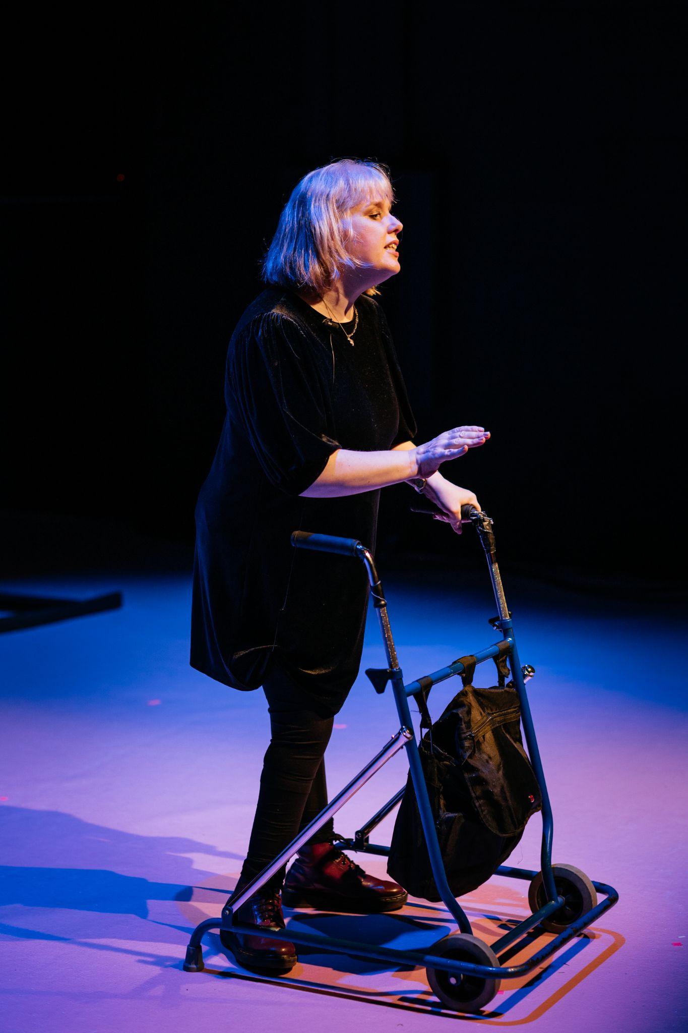 Artist Mairéad Folan stands on a stage lit in purple and pink hues. She is dressed all in black and leans on a black walking frame. They face forward, speaking to an audience who are out of frame, and gesturing with their right hand. Photo by Simon Lazewski.