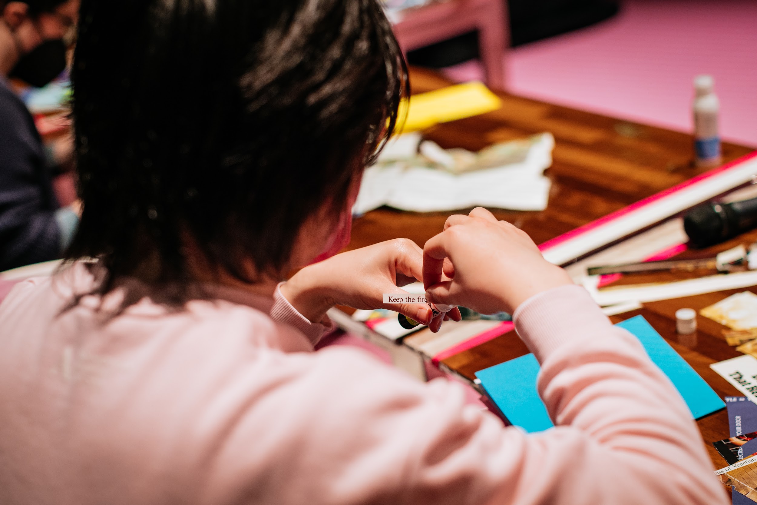 A figure in pink at a zine workshop, captured from behind. Their hands pick up a small magazine cutting, a line of text reads “Keep the fire”. Photo by Simon Lazewski.