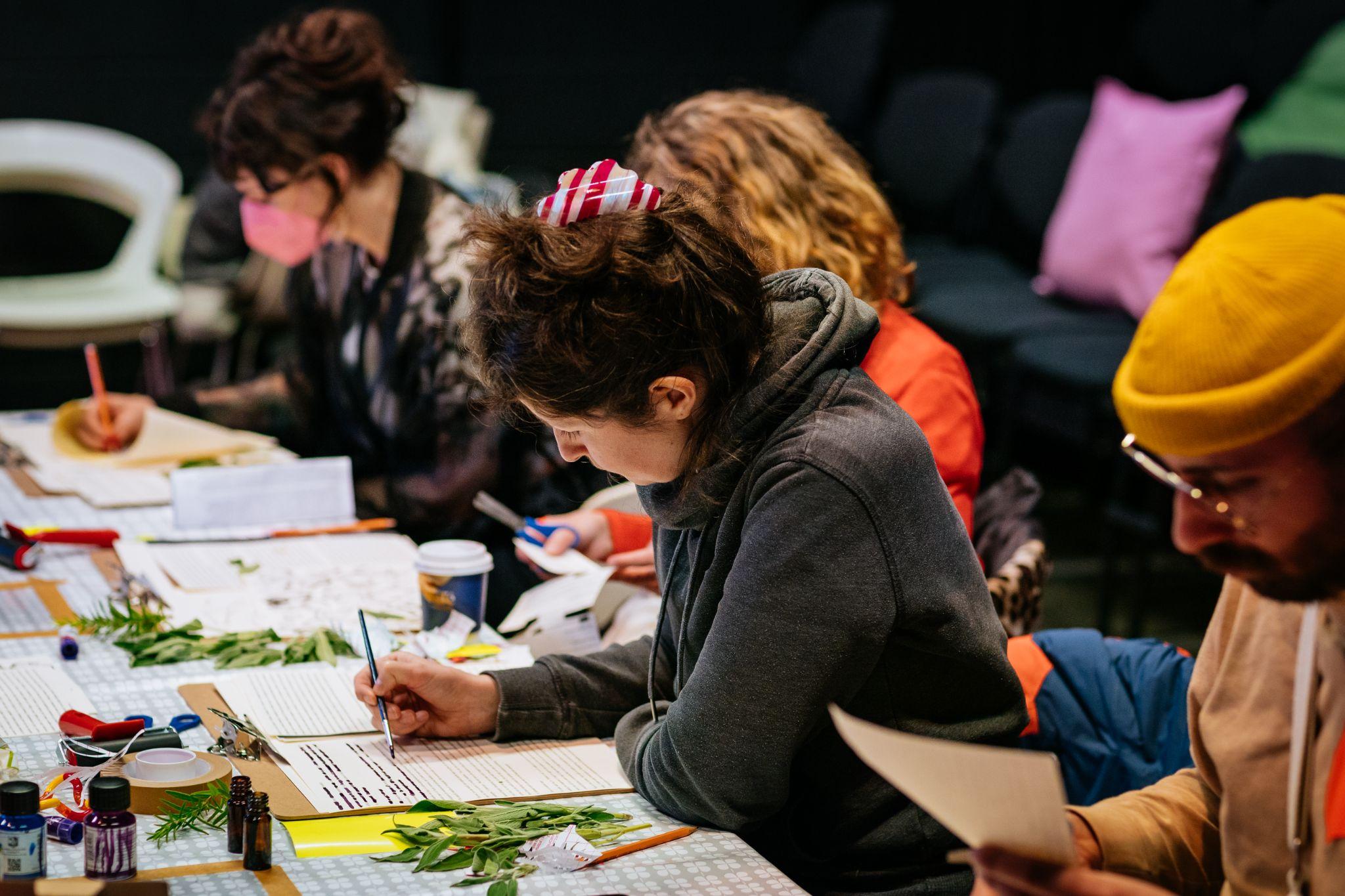 An image taken from the side shows four people sitting at two desks positioned at an angle to each other in a workshop setting. They are all dressed in various colours and have different hair shades, and one wears a yellow hat. They appear to be cutting paper with scissors, drawing and painting. Various materials, including more texts, small boxes, leaves, and paints, are on the table in front of them. Photo by Simon Lazewski.