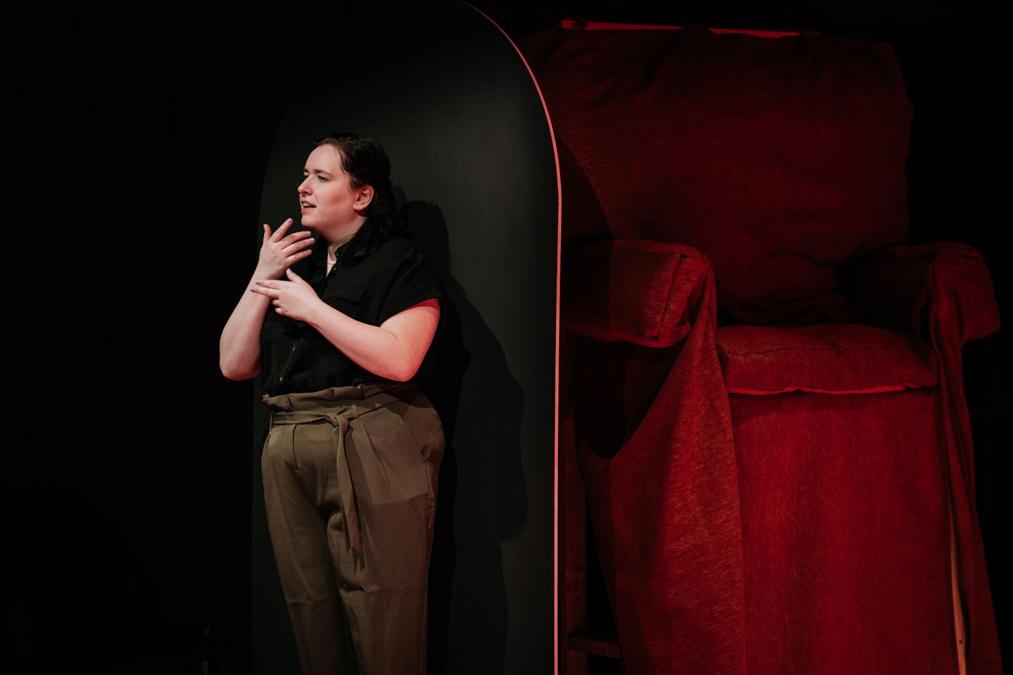 A person with pale skin and long dark hair tied back stands against a dark background, angled away from the camera. They are an ISL sign language interpreter. They wear a black top and green trousers, engaged in sign language. Behind them, a large dimly lit red chair is partially visible behind a black screen. Photo by Simon Lazewski.