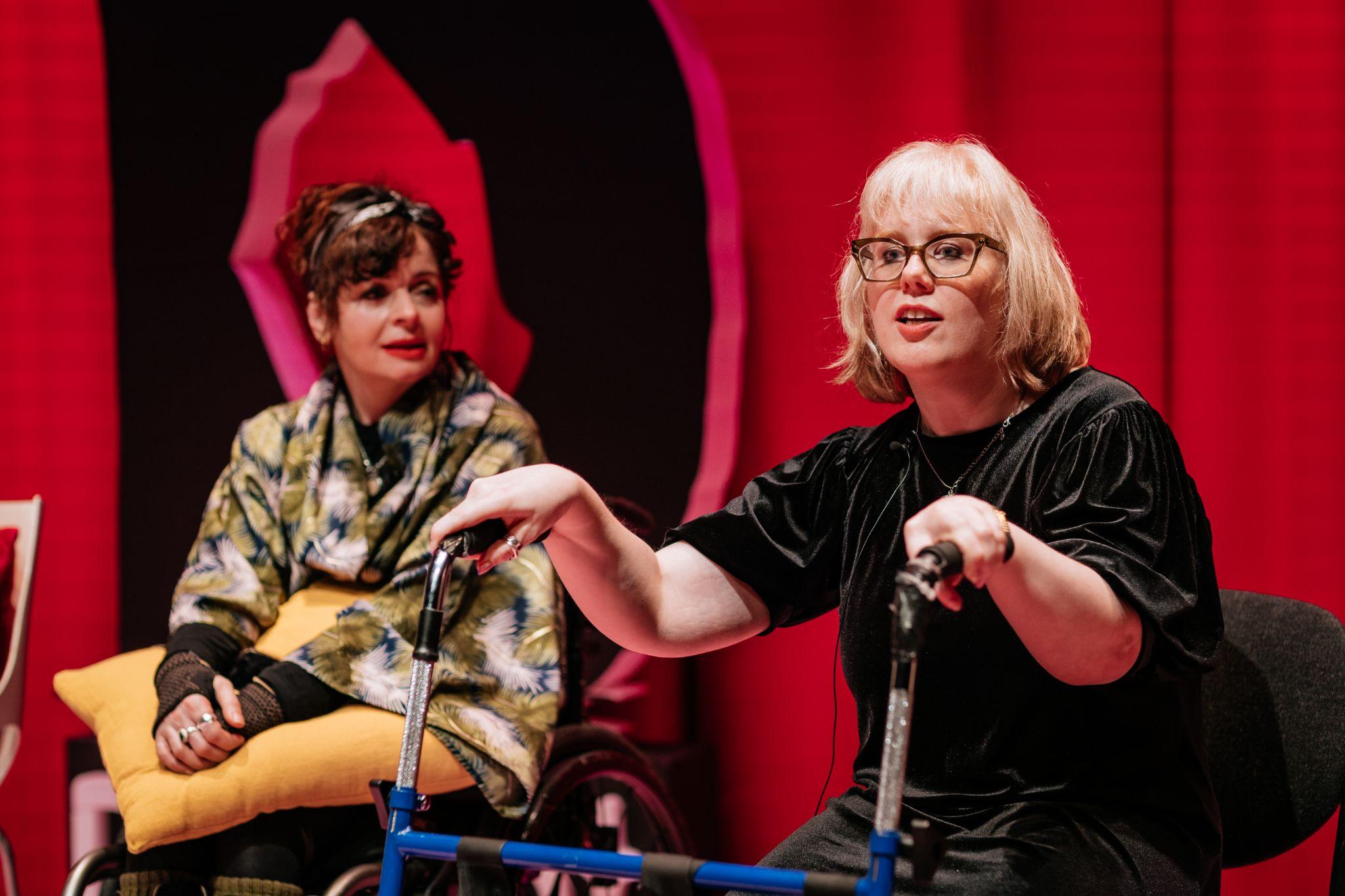 Two people sit facing forward at a slight angle to the camera. One person, with light shoulder-length hair and glasses, wears a black top and rests their hands on a walking frame, looking forward. The other person, with dark hair and light skin, looks towards the first person while resting their hands on a yellow pillow. Behind them is a vibrant pink background with a large black letter “D” for Disrupt Disability Arts Festival. Photo by Simon Lazewski.