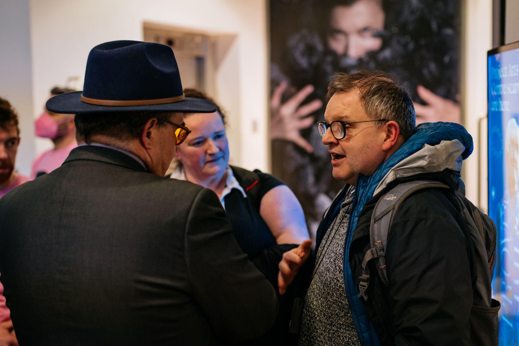 Two individuals converse in an art exhibition, facing each other at an angle to the camera. The room is filled with people and artwork displayed on the walls. Photo by Simon Lazewski.