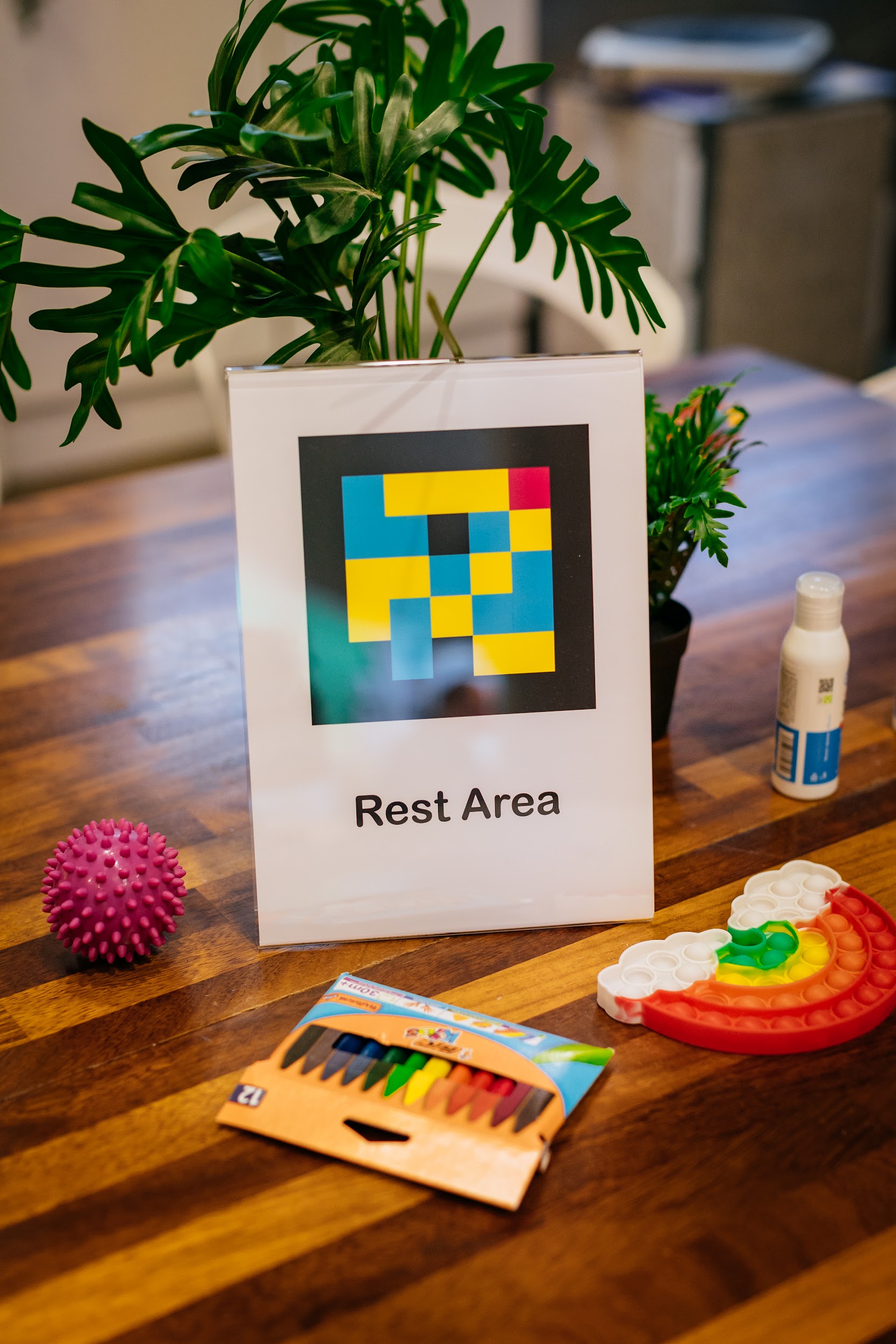 A collection of items on a tabletop. A sign beside a plant reads “Rest Area”. It has a Navi code on it. Other items on the table include a bottle of hand sanitiser, crayons and various stim toys. Photo by Simon Lazewski.