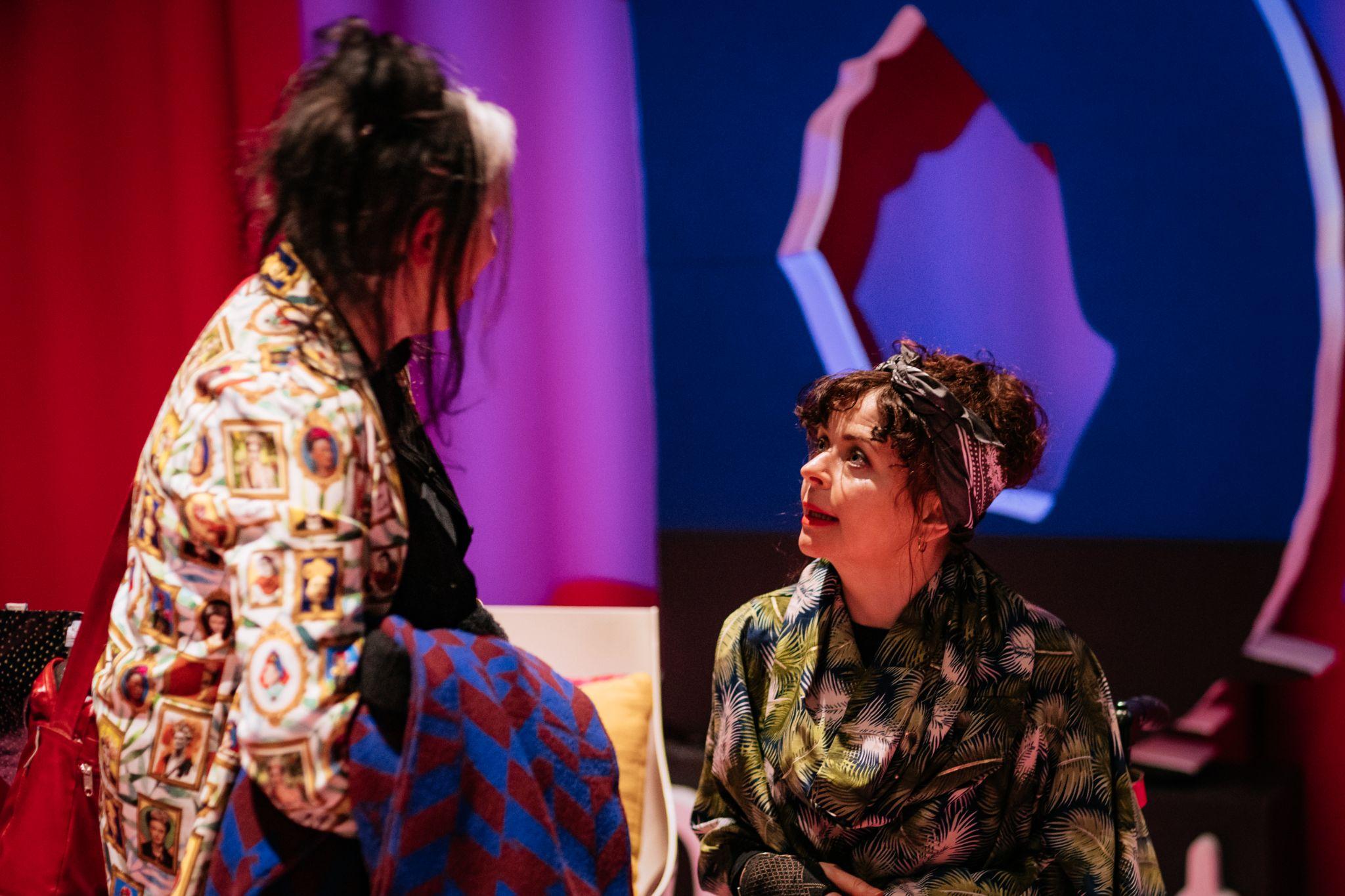 Two individuals with long dark hair tied up engage in conversation, wearing brightly coloured tops with various patterns. Behind them, a vibrant pink and purple background showcases a large blue and pink letter “D” for Disrupt Disability Arts Festival. Photo by Simon Lazewski.