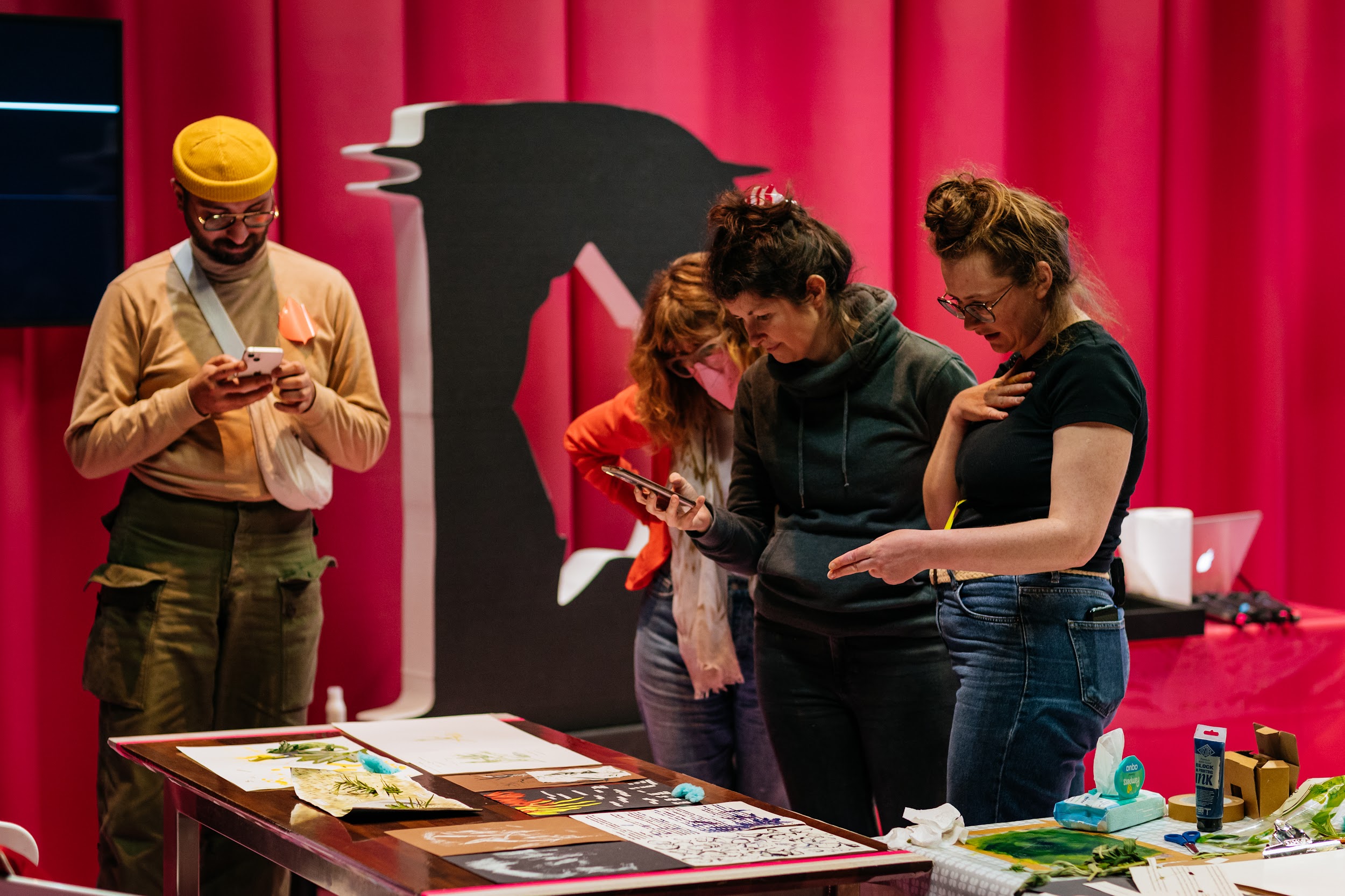 Artist Michelle Hall reviews work with three participants at a workshop. They are surrounded by art materials. Behind them is a large styrofoam D, the disrupt logo. They are in front of a pink curtain. Photo by Simon Lazewski.