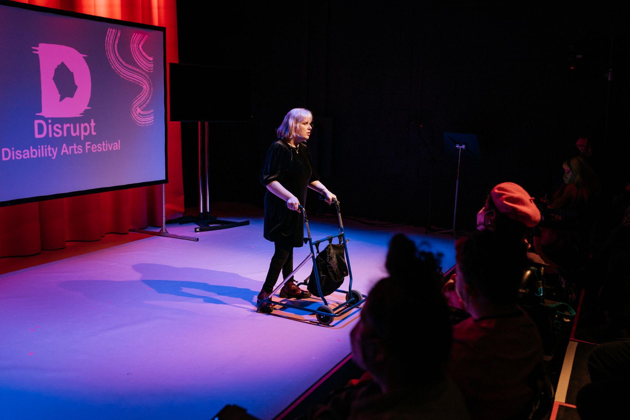 A fair-haired individual with shoulder-length hair and pale skin stands on a lit stage, wearing dark coloured clothing and with a black walking frame. They engage with an audience, who are mostly unseen. The stage is lit in purple and pink hues. Viewed from a diagonal perspective within the audience area, silhouetted spectators sit facing the stage. Behind the individual, there is a projection screen on a stand displaying the logo for Disrupt Disability Arts Festival. Photo by Simon Lazewski.