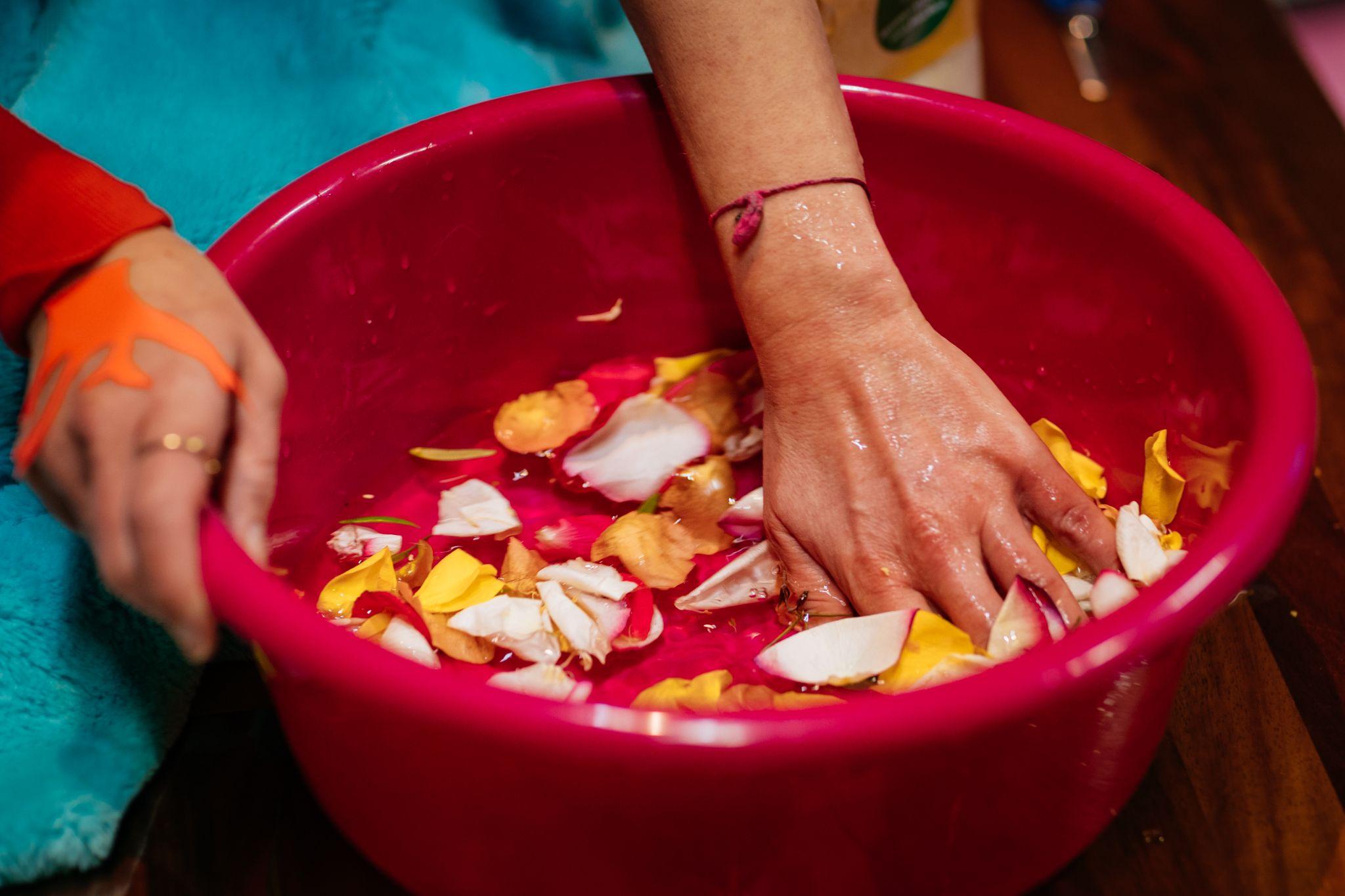 The image shows a pair of light-skinned hands in a pink basin containing water. In the water petals are floating. The petals are pink, yellow, and white. Photo by Simon Lazewski.