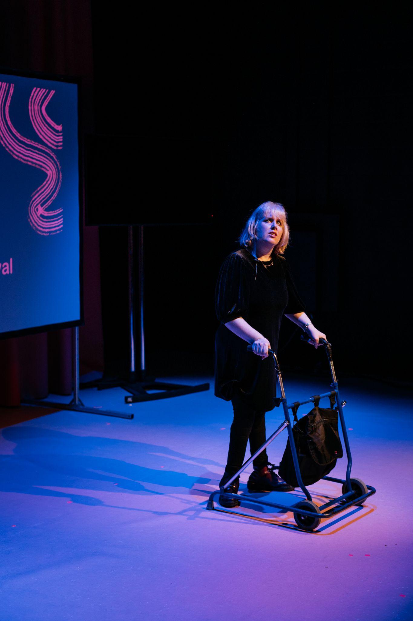 Artist Mairéad Folan stands on a stage lit in purple and pink hues. They gaze upward quizzically. They are dressed in black and lean on a walking aid. In the background, there is a projection screen on a stand displaying the logo for Disrupt Disability Arts Festival, mostly out of the frame. Photo by Simon Lazewski.