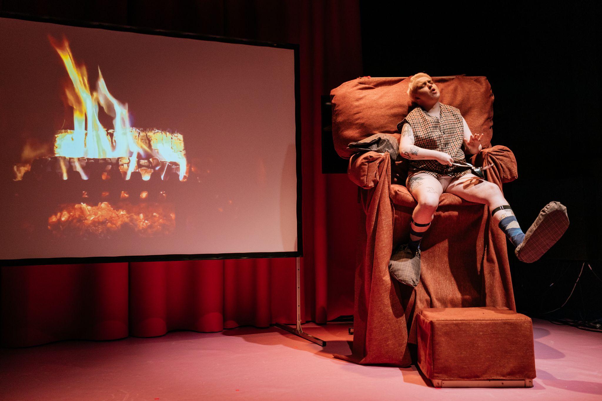 Them Fatale, a character, sits on a comically oversized orange chair with a footstool. They are wearing a brown, orange, and cream tartan plaid baggy waistcoat, with black straps around their lower legs, and oversized slippers in similar colours and material. Their legs are stretched in opposite directions. To their right, a projection screen displays an image of a fire. Photo by Simon Lazewski.