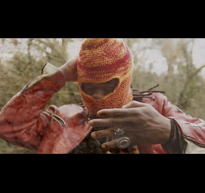 This is an image of artist Tobi Balogun’s performance. It is a still of a video art piece Tobi created for Disrupt. In the center , we see Tobi who is a black Yoruba-Irish male. He is dressed in a red leather jacket and is wearing a woollen knitted headpiece in red and yellows on his head, only revealing his eyes and part of his nose. He is looking directly at the camera, with one hand placed behind his head and one hand in the foreground of the photo, showing the back of his hand. On his hand he wears silver ring jewellery. The background is a green woodland. Photo by Simon Lazewski.