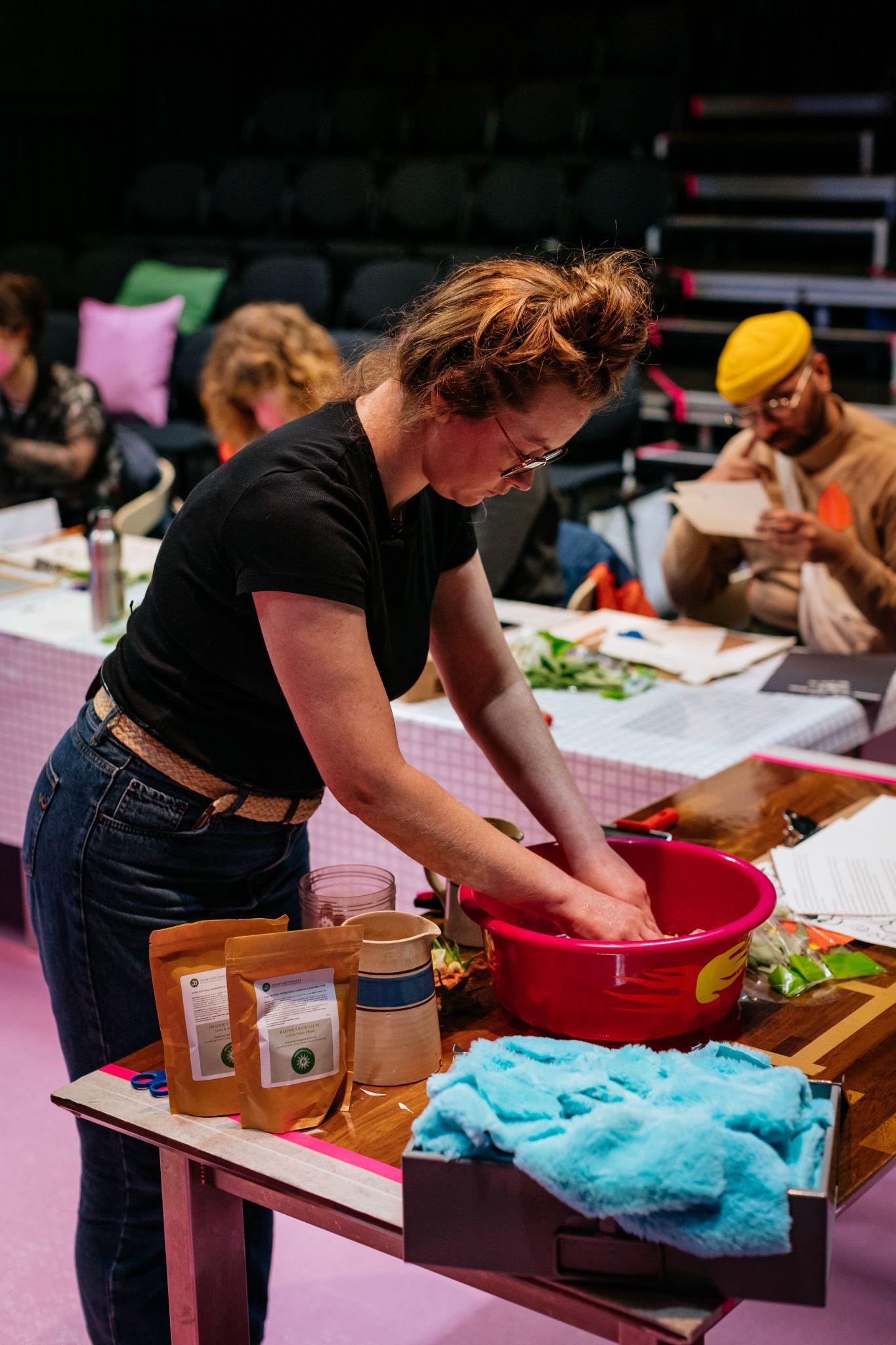 An image of a person standing at an angle to the camera in the foreground. They have pale skin and long auburn hair tied in a knot. They wear glasses, a black top, and jeans. The person is leaning over with both hands in a pink basin placed on a table. Other objects on the table include a jug, brown paper packets, and blue fluffy material. In the background, a group of people are out of focus seated at a desk, engaged in craft-like activities. Photo by Simon Lazewski.