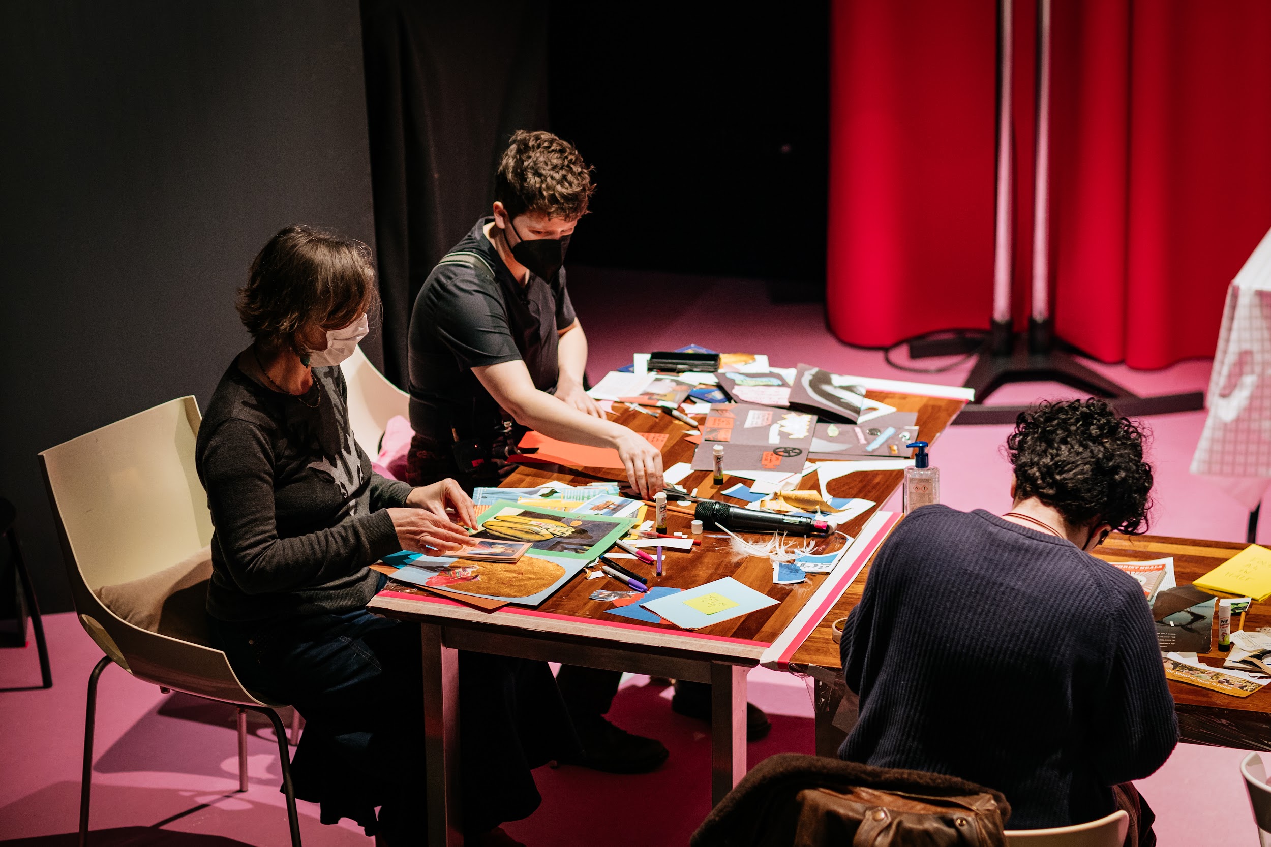 Two participants of a workshop sit with artist Ailing Reina. They wear facemasks. The workshop takes place on the Disrupt festival stage, where the carpet and stage curtain are pink. The tabletops are full of magazine cuttings, coloured paper and glue sticks. Aisling and the participants are very focused on their work. Photo by Simon Lazewski.