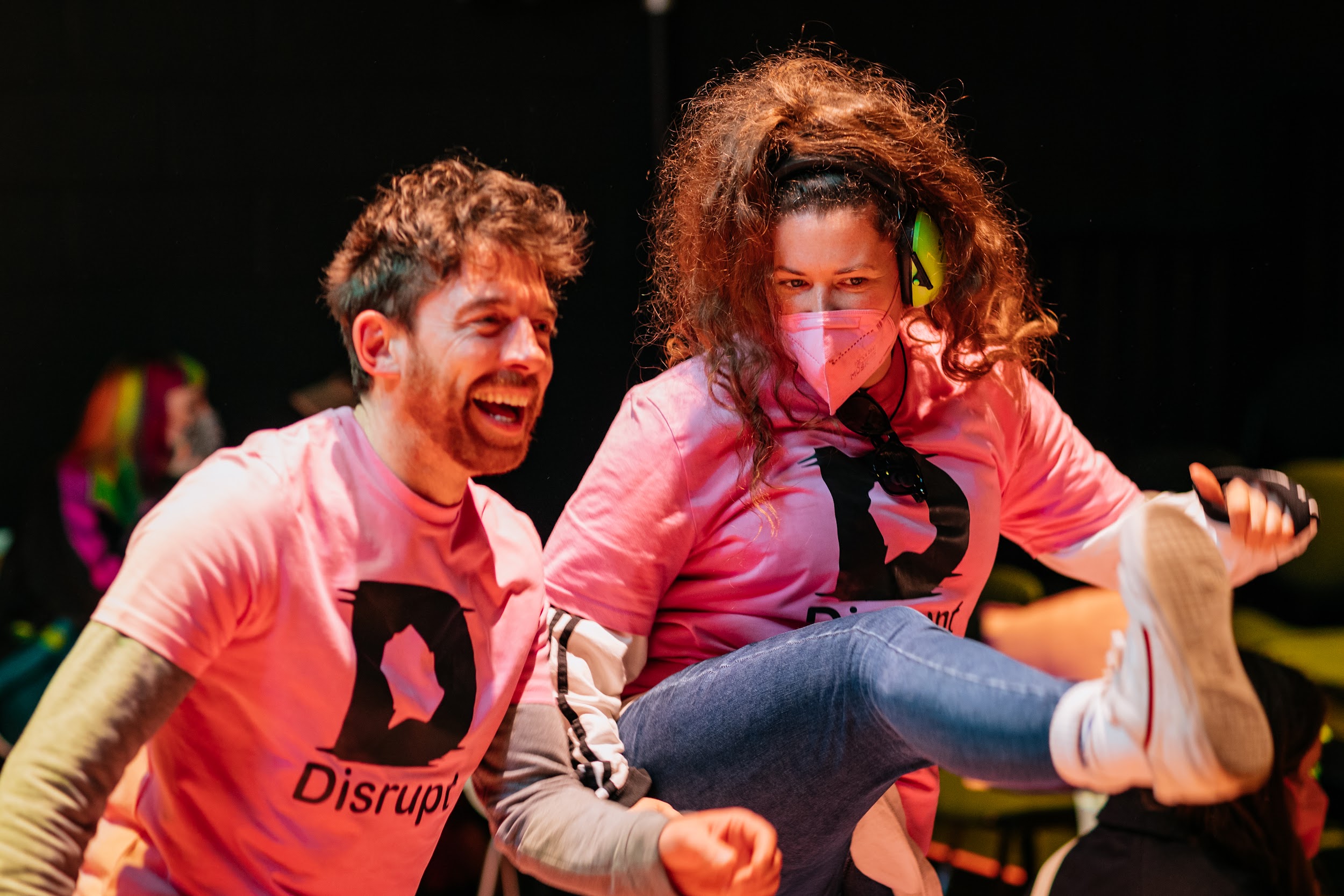 Festival director Alan James Burns dances with a crew member at the disconnect closing party. Alan has short brown hair and wears a pink disrupt t-shirt. The other person has long brown hair worn in a wild ponytail. She wears a pink t-shirt and facemask. She is kicking her leg high in the hair.