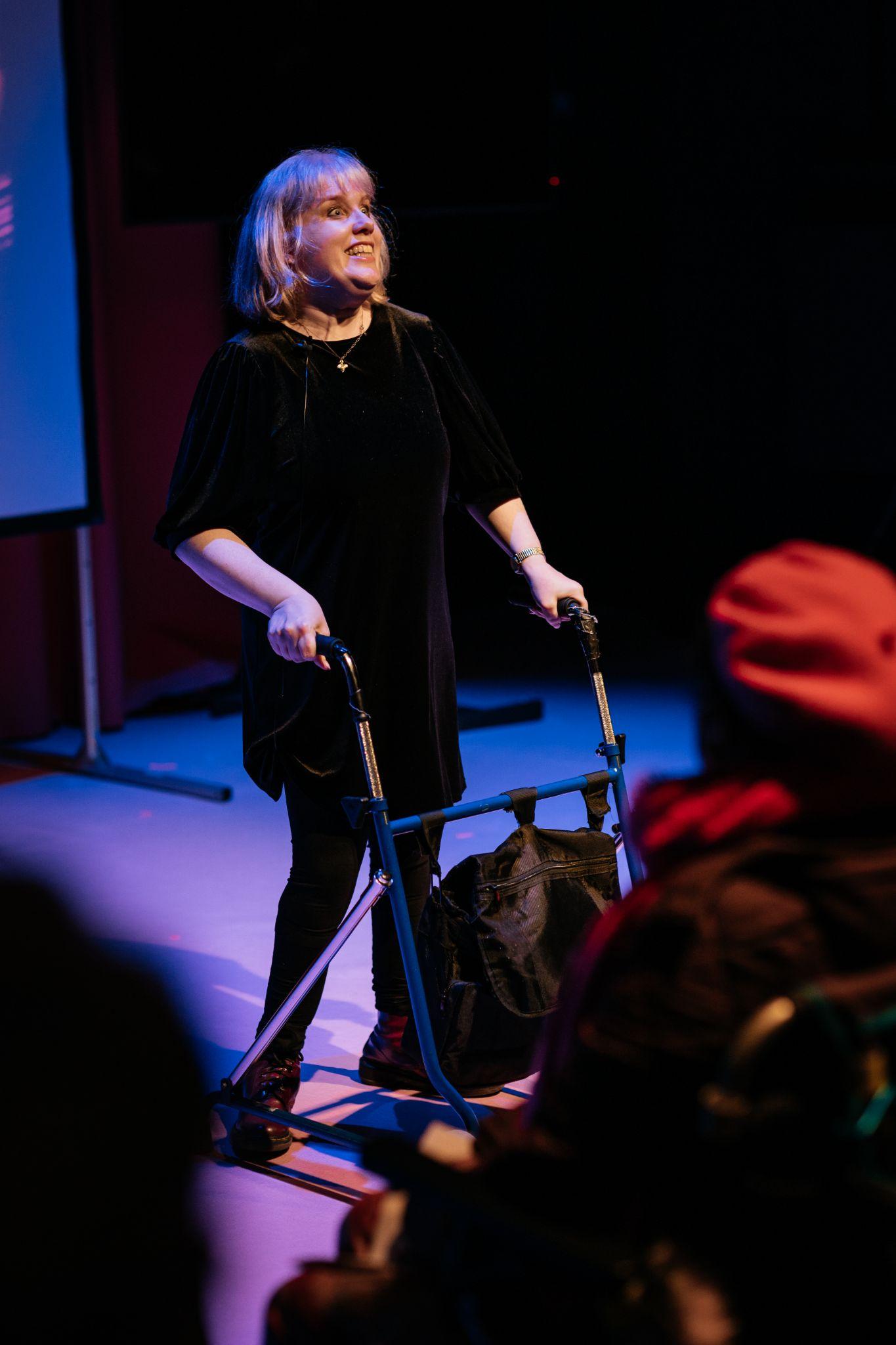 fair-haired individual with shoulder-length hair and pale skin stands on a lit stage, wearing dark coloured clothing and with a black walking frame. They are smiling as they engage with an audience, who are mostly unseen. The stage is lit in purple and pink hues. Viewed from a diagonal perspective within the audience area, silhouetted spectators sit facing the stage. In the background, a small corner of a projection screen can be seen. Photo by Simon Lazewski.