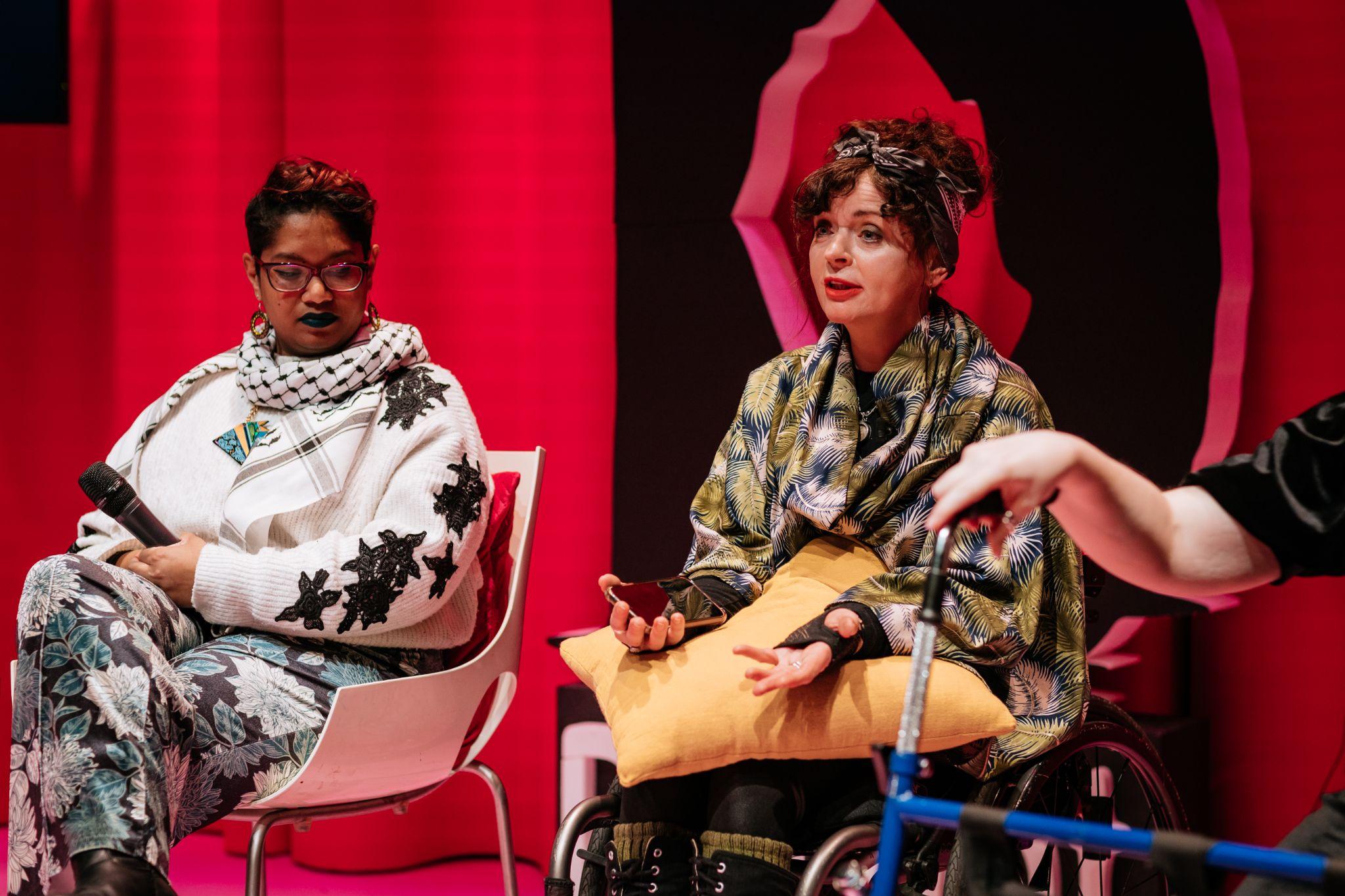 Two people sit facing forward. One person, with dark hair and light skin, speaks to an unseen audience while holding a yellow pillow. The other person, with short dark hair and dark skin, holds a microphone and looks down. Behind them is a vibrant pink background with a large black letter “D” for Disrupt Disability Arts Festival. A hand rests on a walking frame to the right. Photo by Simon Lazewski.