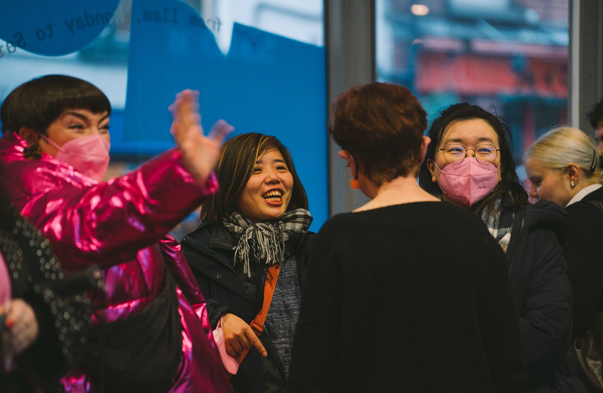 A group of people standing in a circle and looking in various directions in a busy space. Two of them wear bright pink masks, all standing in front of a window. They look excited, and one person dressed in a bright neon pink shiny jacket gestures with their right hand. Photo by Leanne Sullivan.